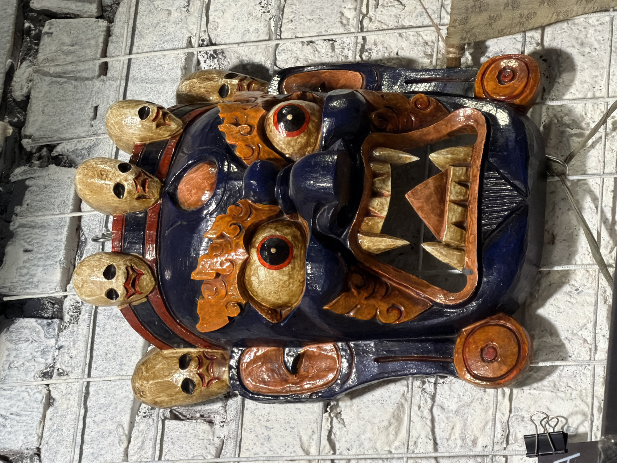A colorfully painted wooden mask is displayed on a white-painted brick wall with a metal grid. The mask is predominantly blue with brown and orange details. It features large, round eyes and a mouth with a triangular pattern. Smaller skulls are attached to the sides of the mask. The scene appears to be indoors. There's no discernible connection to Moscow, Russia in the image.