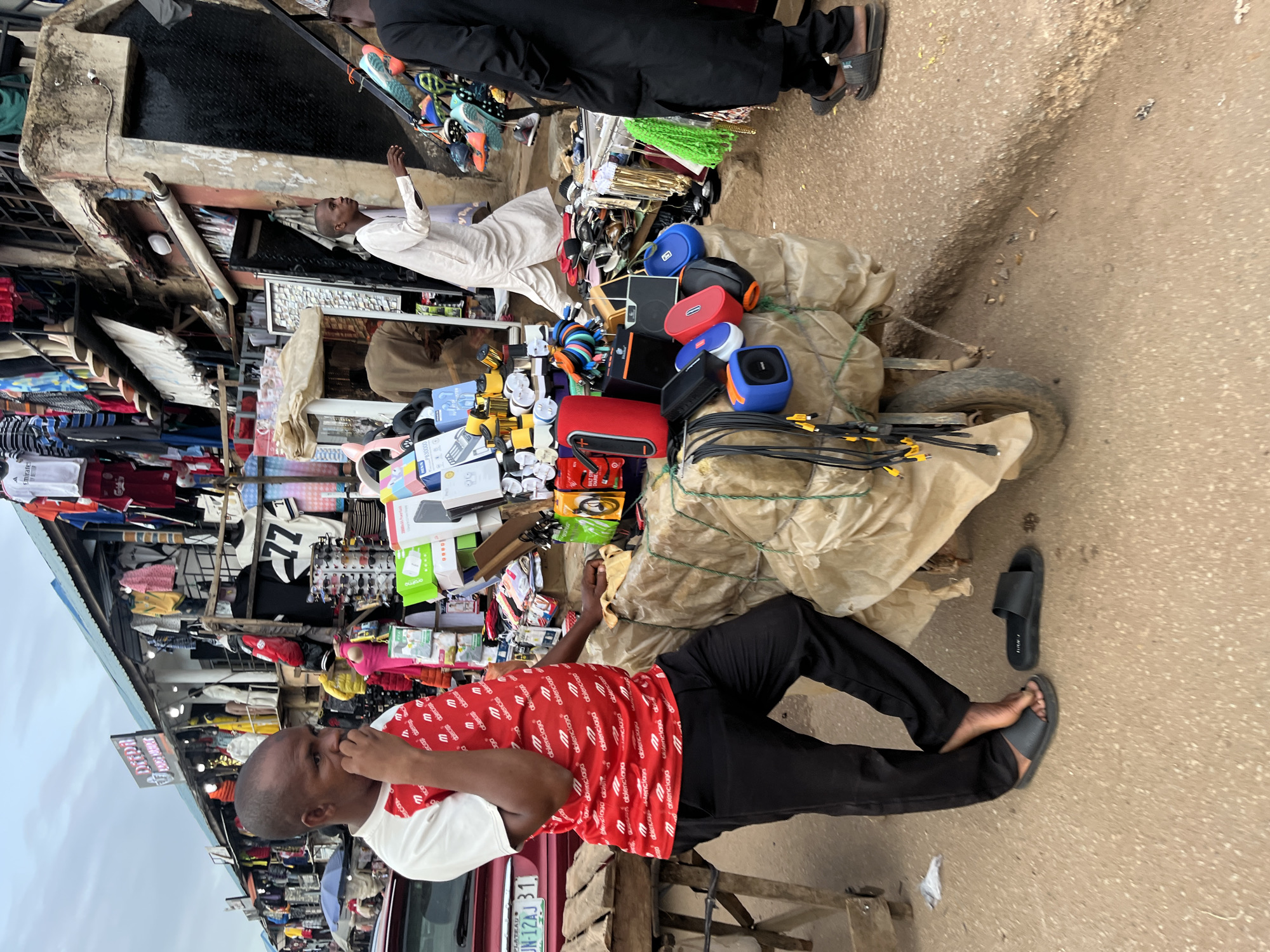 In Bukuru, Nigeria, a man stands beside a cart laden with consumer electronics and wrapped goods. He wears a red and white t-shirt with a repeating logo and black trousers, with one foot bare in a black sandal and the other in a black slide sandal. The cart, partially covered with tan sheeting, displays various items including portable speakers, cables, and what appear to be packaged electronic devices. Behind him, a bustling marketplace is visible with stalls selling clothing and other merchandise. A red car is parked to the left, and other individuals are seen in the background, engaged in shopping or traversing the crowded street. The ground is paved with asphalt and dirt, with scattered debris suggesting a busy urban environment.