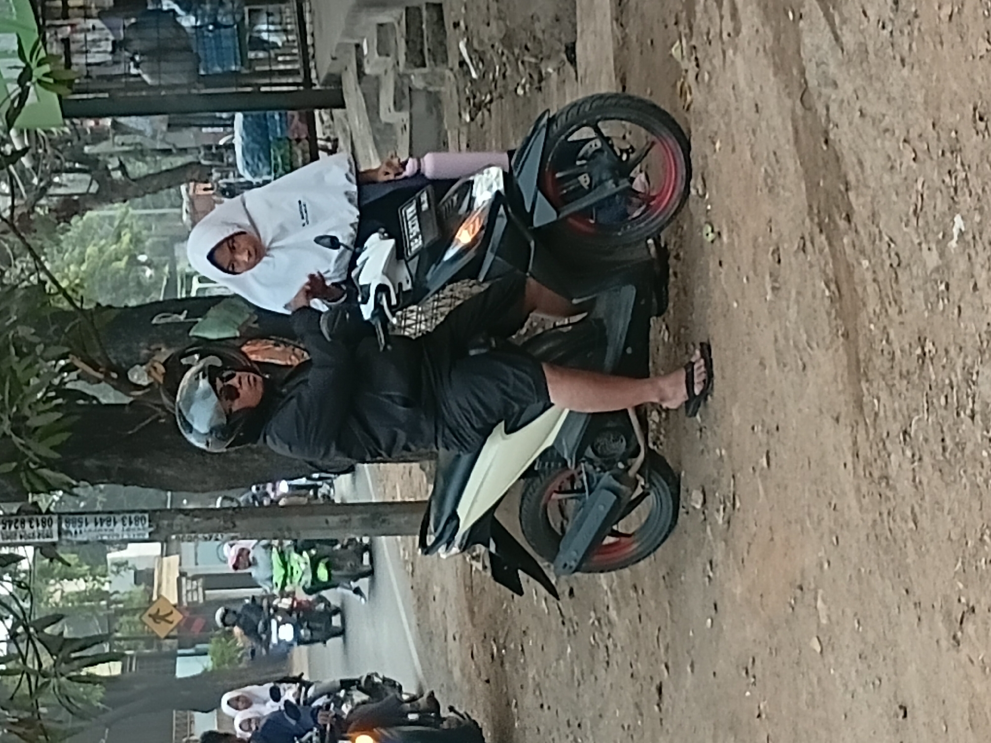 In Bekasi, Indonesia, two individuals are positioned on a black and white motorcycle. A person in a black jacket and sunglasses, wearing a helmet, is operating the vehicle. Beside them, a person in a white hijab is seated. The motorcycle is on a dirt road, adjacent to a paved road with other vehicles, including motorcycles, visible in the background. The scene appears to be a street in a moderately populated urban area.