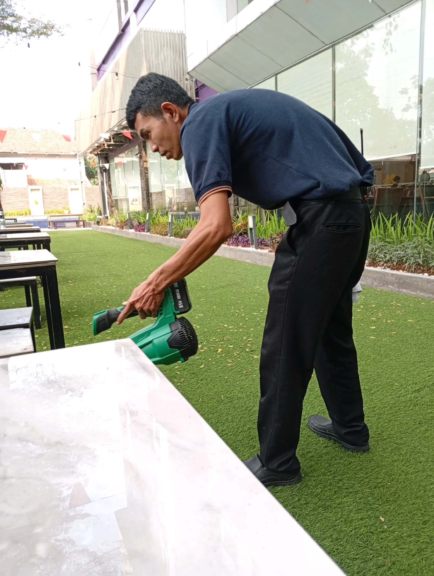 In Binong, Indonesia, a man is using a green leaf blower to clean a white, marbled table. He is wearing a dark blue polo shirt and black pants. The table is situated on a bright green, artificial turf lawn. In the background, there are several other tables and chairs, as well as a building with a modern design, featuring glass windows and a purple accent. The overall scene suggests an outdoor seating area, possibly at a restaurant or cafe.