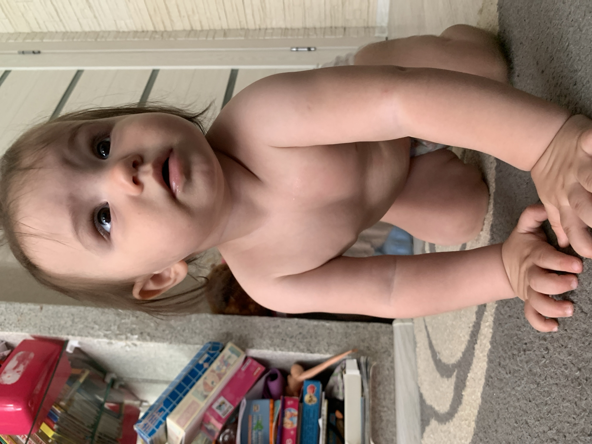 The image shows a close-up, eye-level view of a baby on its hands and knees on a carpet. The baby is facing slightly to the right, with its head tilted upwards and to the right, looking at something out of frame. The baby has dark hair, brown eyes, and fair skin. Its mouth is slightly open, with a bit of moisture visible on its lower lip. The baby is not wearing any clothes except for a diaper.
The background is out of focus, but appears to be an indoor setting. To the left, there is a shelf with various items on it, including what appear to be children's books or boxes, some of which are brightly colored. A portion of a wall with horizontal lines is visible behind the baby. To the right, a door frame is visible. The carpet is grey with a cream-colored circular pattern.
The scene is consistent with a domestic setting, likely a child's room or play area. There are no indications of any specific location within the city of Chaikovsky, Russia, beyond the general assumption of an indoor residential environment. The lighting suggests it is daytime. The focus is on the baby, with the surroundings blurred to emphasize the subject.
