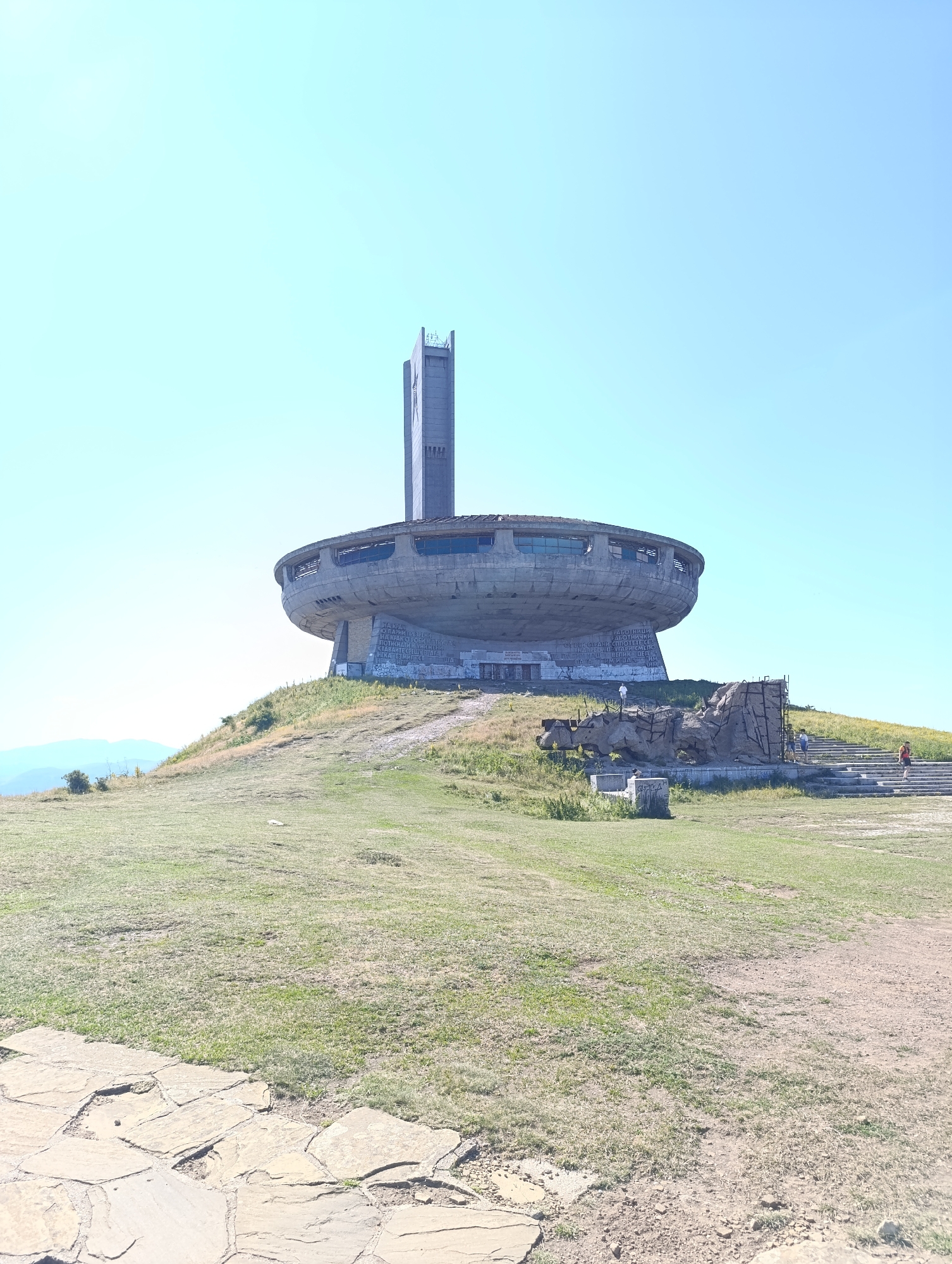 The image depicts the Buzludzha monument, a concrete, saucer-shaped structure with a tall tower, situated on a grassy hilltop. The monument is located in the vicinity of Shipka, Bulgaria. The sky is clear and blue. The monument shows signs of decay and graffiti. Several people are visible on the grounds, including some walking up a stone staircase on the right side of the monument, and others near the base. The foreground features a path paved with irregular stone slabs. The surrounding environment is a grassy, undulating landscape with some sparse vegetation.