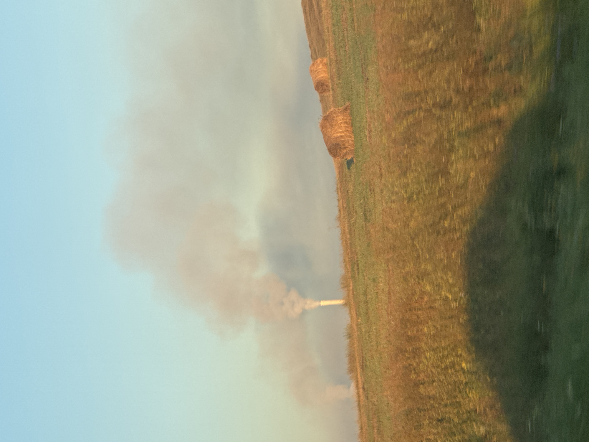 This is a photograph of a landscape with industrial emissions and agricultural elements, likely taken in or near Magnitogorsk, Russia. In the background, a tall, slender industrial chimney is emitting a thick plume of light-colored smoke that drifts across the sky. To the left of this, another less distinct industrial emission is visible, also in the form of a smoke plume. The sky is a clear, pale blue, with the sun casting a warm, golden light on the scene. In the mid-ground and foreground, a grassy field is illuminated by this light, with several large, round hay bales scattered across the sloping terrain. The texture of the grass appears dry and golden, suggesting it may be late summer or early autumn. The bottom portion of the image is blurred, indicating motion, possibly from the viewpoint of a moving vehicle, with green vegetation visible in the very foreground. The overall impression is one of an industrial area interacting with a rural or semi-rural environment.