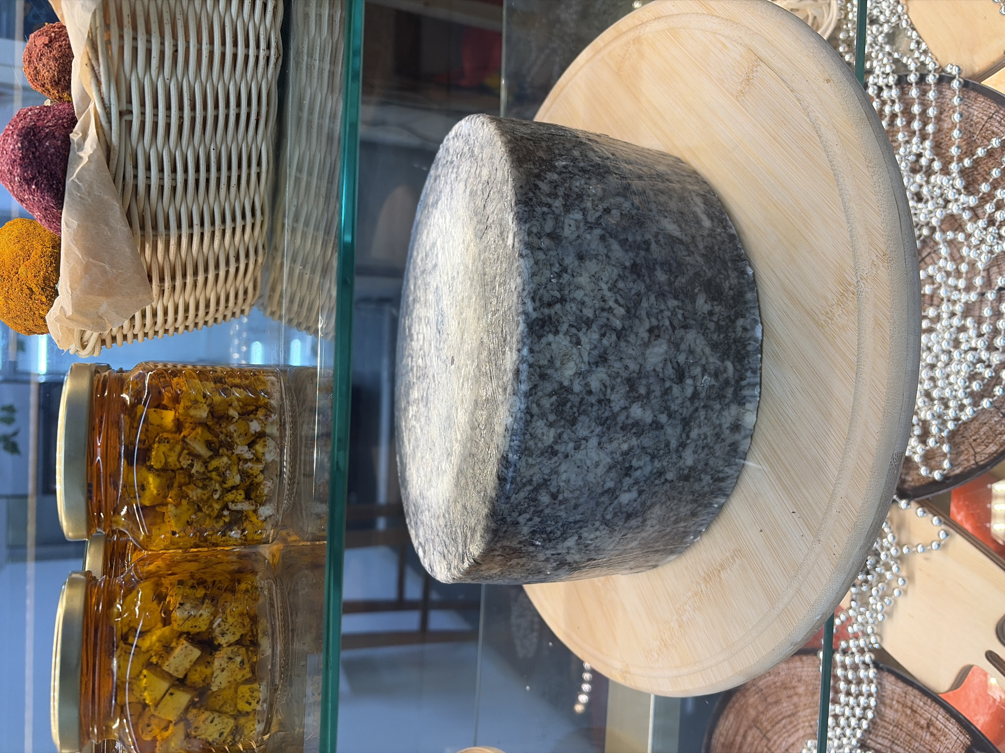 In a refrigerated display case in Kazan, Russia, a large wheel of marbled grey cheese sits on a light-colored wooden cutting board. The cheese is covered in plastic wrap and appears to be a hard variety. Behind the cheese, to the left, are two glass jars filled with cubed food items submerged in a golden liquid, possibly pickled vegetables or cheese. To the right of the jars, a woven basket contains three round, brightly colored food items, possibly truffles or cheese balls coated in spices. The display case has a glass shelf, and the background shows reflections of furniture. The overall setting suggests a food market or a specialty cheese shop.