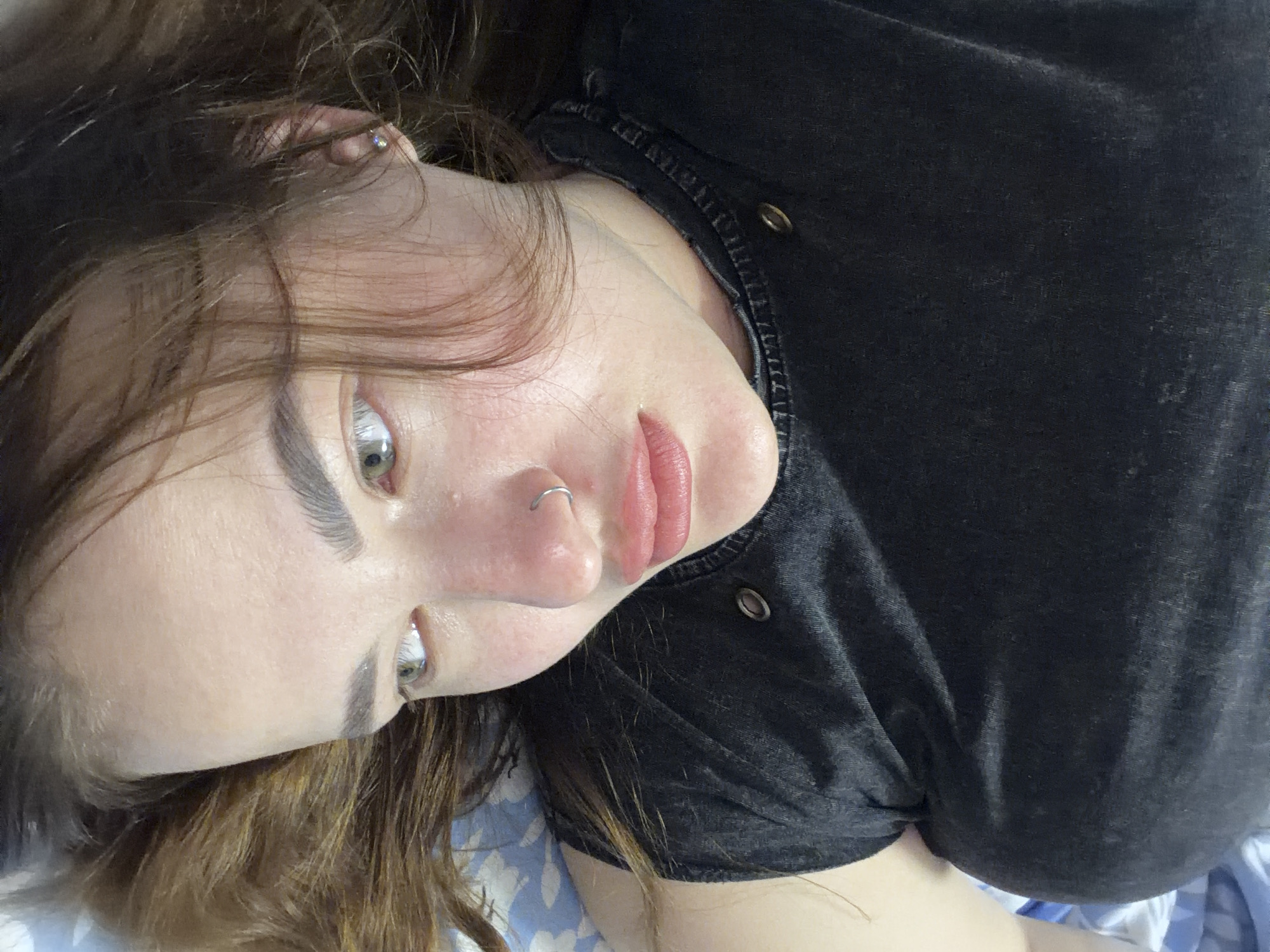 A woman with light skin, light brown hair, and hazel eyes is visible in a close-up, downward angle shot. She has a nose ring in her left nostril and a stud earring in her left ear. She is wearing a dark-colored, collared shirt with button details. The background shows a floral patterned surface. There are no indications this picture was taken in Krasnodar, Russia.