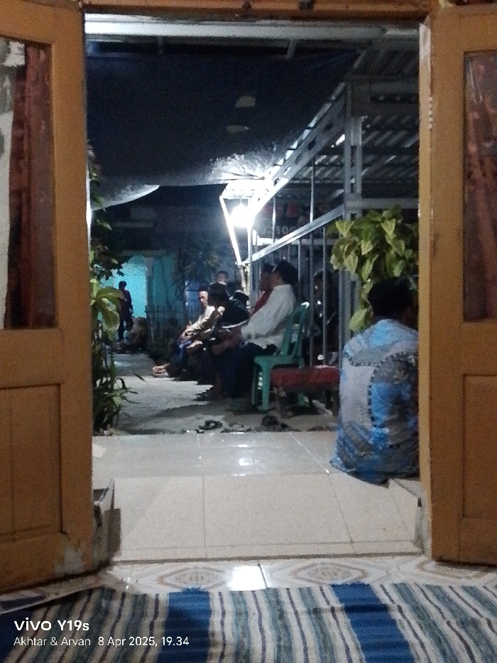 The image shows a scene taken from inside a building in Gegesik, Indonesia (latitude: -6.538, longitude: 108.43). It appears to be a residential area, possibly a home. Several people, mostly adults, are seated in a covered area outside the building. There are chairs present and the people are engaged in conversation. The environment is dimly lit with a single light source illuminating the scene. The ground is tiled and a blue and white striped mat can be seen in the foreground. The image appears to be taken through an open doorway, with the camera positioned to capture the scene from inside the building. The doorway is wooden and partially open, allowing the viewer to look out into the outdoor gathering space. There is a lot of greenery present. The scene gives the impression of a casual, informal gathering. The people are sitting in a relaxed manner and talking to each other. The image was taken in the evening as it is dark outside.