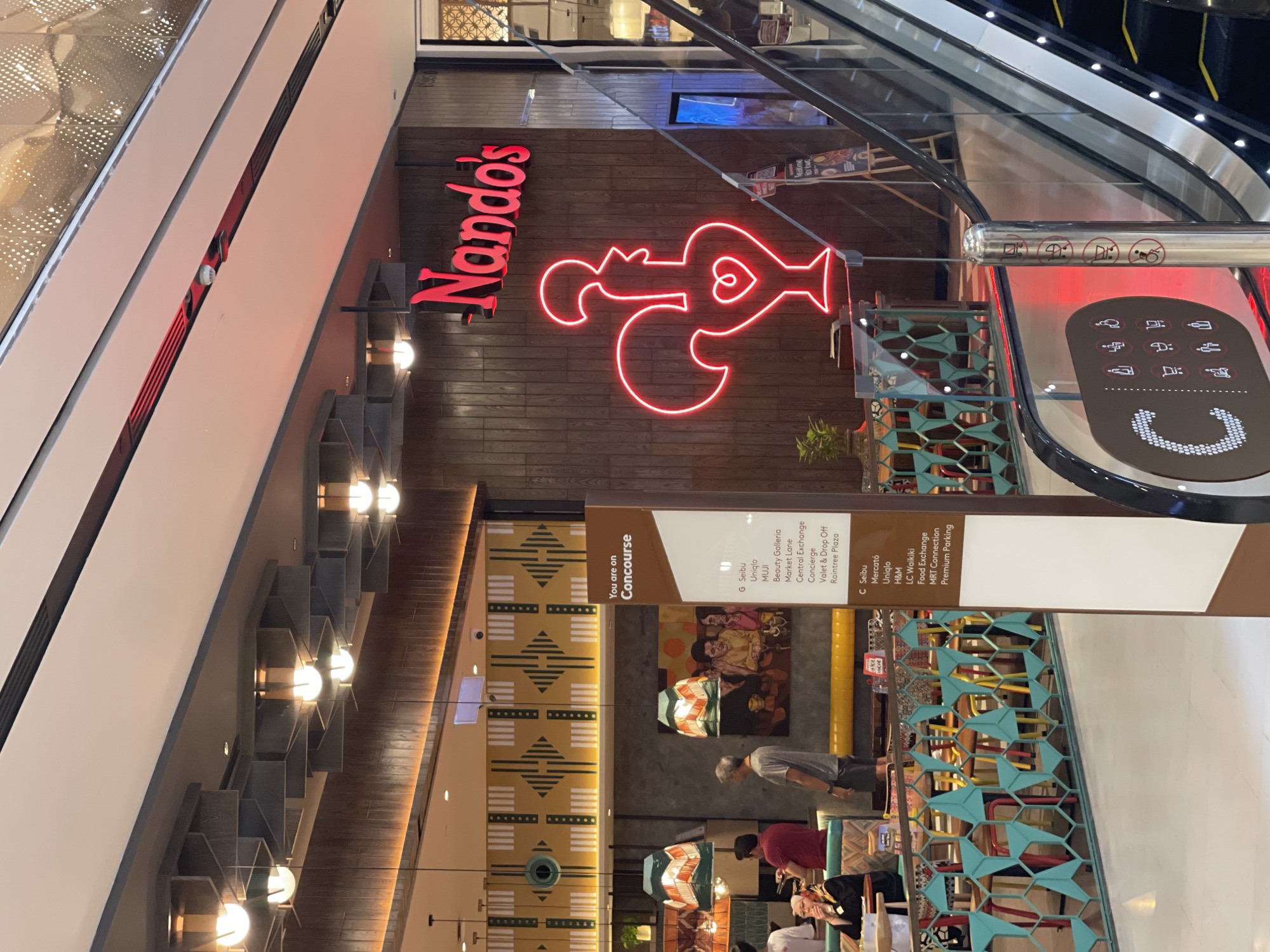 The image shows the exterior of a Nando's restaurant, illuminated by a large red neon sign of their iconic rooster logo and the Nando's text logo. The restaurant is situated within a modern shopping mall environment, indicated by the presence of an escalator with informational signage. The signage identifies "You are on Concourse" and lists various shops and services such as "Seibu", "Uniqlo", "MUJI", and "MRT Connection," suggesting the mall is Pavilion Kuala Lumpur, a well-known shopping destination in Kuala Lumpur, Malaysia. The interior of the restaurant is partially visible, featuring wooden paneling and decorative lighting fixtures. Several people are seen in the background, some seated at tables and others standing, suggesting the restaurant is open for business. The overall scene captures a typical day at a busy shopping mall in Kuala Lumpur.