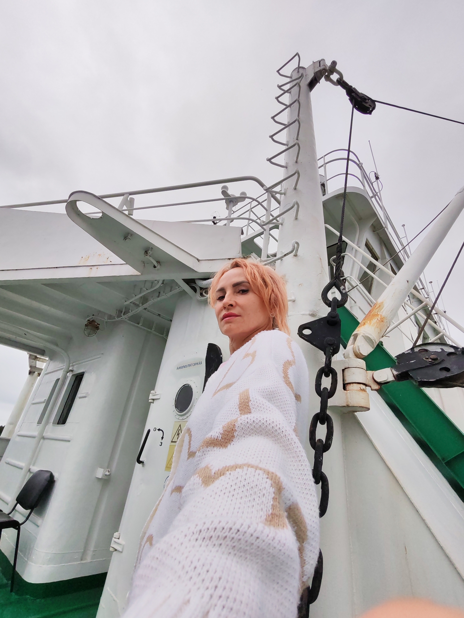 A woman with short, pink hair and light-colored makeup is posing for a selfie on the deck of a white ship. She is wearing a white knitted sweater with beige abstract patterns. Behind her, the ship's structure is visible, including a large white mast with a ladder, rigging, and metal chains. The sky is overcast and gray. The scene appears to be in an industrial or maritime setting, possibly a port in Shal'skii, Russia, indicated by the Cyrillic text "АККУМУЛЯТОРНАЯ" (Accumulator) on a door. The angle of the shot is from a low perspective, looking up at the woman and the ship's structures.