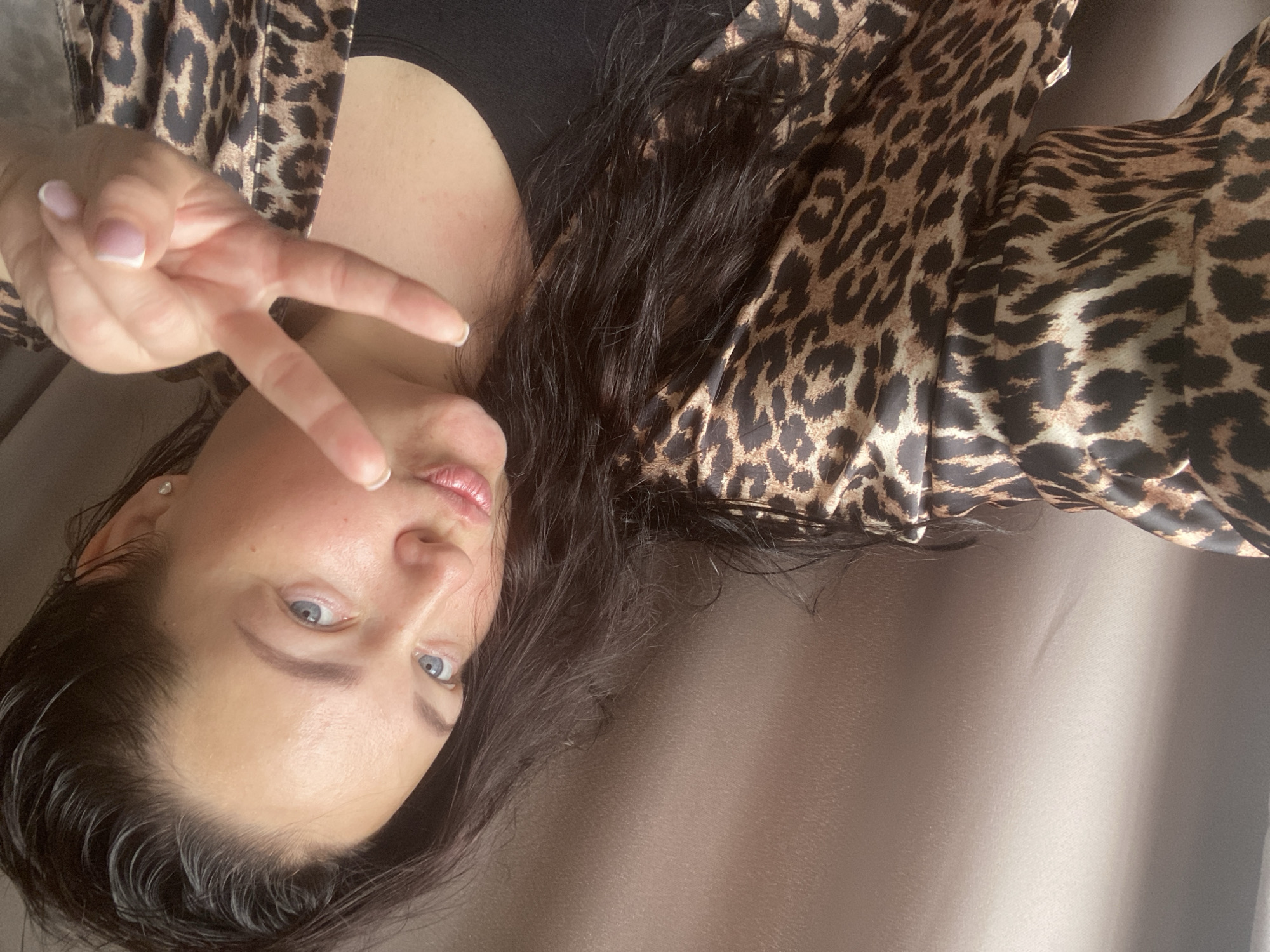 A woman with light skin, blue eyes, and dark hair is posing indoors. She is wearing a black top under a leopard-print robe. Her hair appears wet, and she is making a peace sign with her left hand. The background is a plain light-brown surface. There is no indication of Sochi, Russia, in the image.