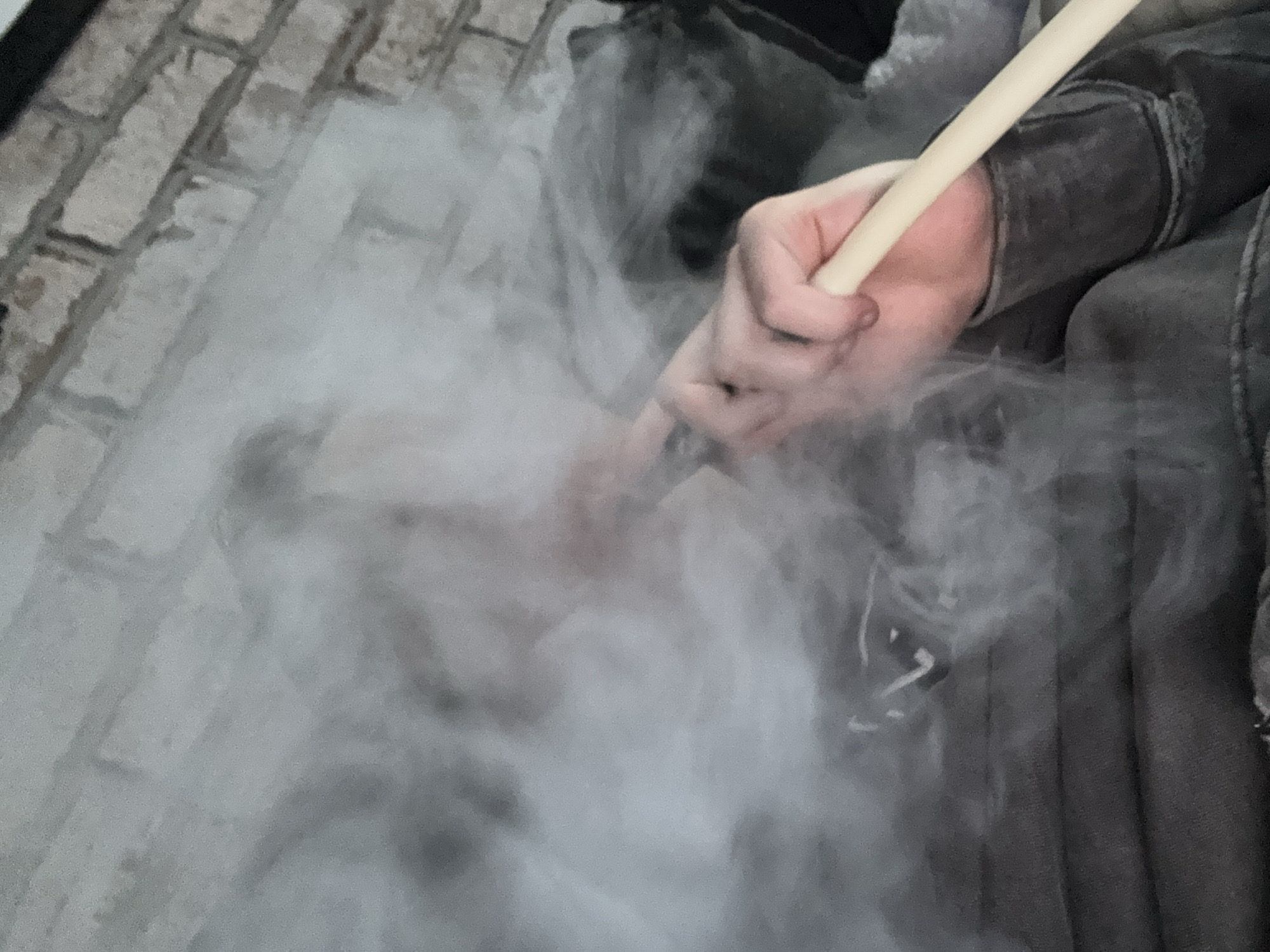 The image shows a close-up of a person's hand holding a hookah hose, with smoke billowing around it. The background appears to be a brick wall, suggesting an indoor or outdoor setting. The lighting is dim, with the smoke being the brightest element in the frame. The person is wearing a dark, textured garment, possibly a jacket or hoodie. The image focuses on the act of smoking a hookah, with the hand and hose being the central subjects. The location in Huixquilucan de Degollado, Mexico, is not explicitly visible in the image itself, but is provided as context.