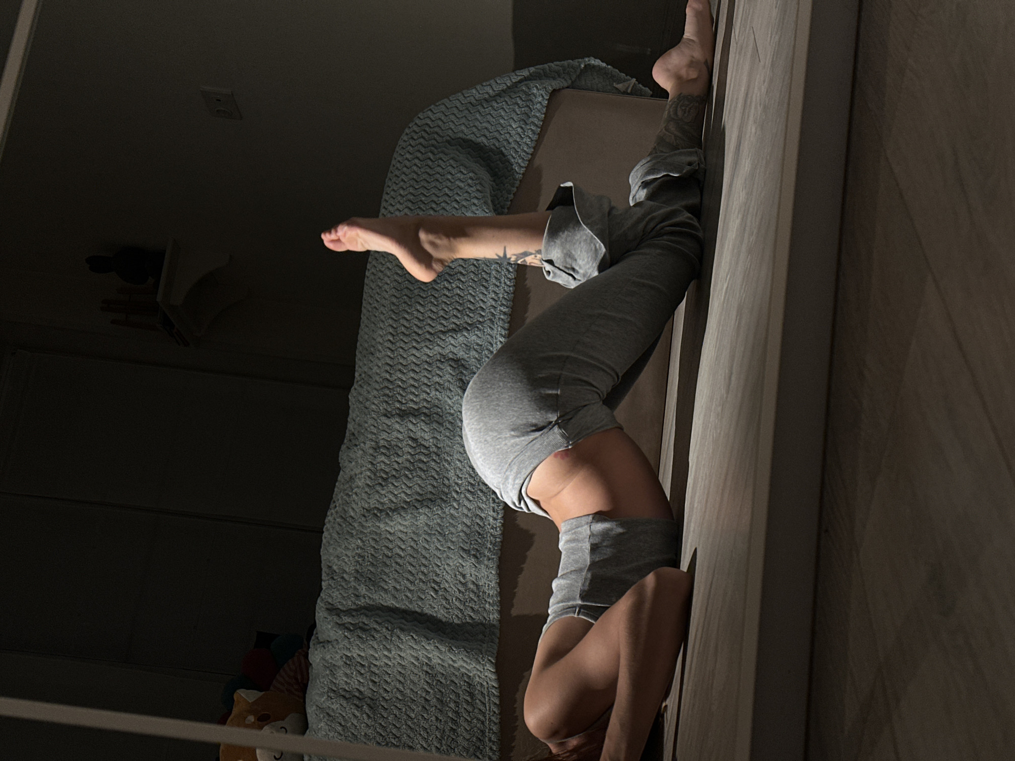 The image depicts a person lying on their stomach on a wooden floor, with their upper body arched backwards. They are wearing grey sweatpants and a matching grey tank top. Their left leg is bent at the knee and extended upwards, while their right leg is extended straight behind them. A bed with a textured blue blanket is visible in the background, along with a shelf on the wall holding some objects. The room is dimly lit, with shadows cast by the low light. There is a reflection in what appears to be a mirror in the foreground. The setting does not provide any information about a specific location in Moscow, Russia.