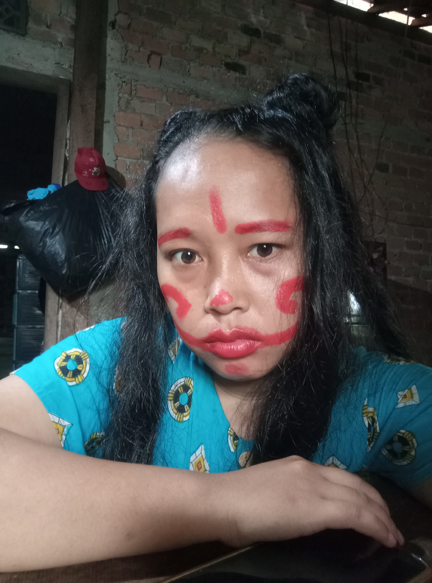 A young woman with black hair styled in two buns and red face paint is looking at the camera in an indoor setting. The face paint consists of red lines on her forehead and cheeks, and a red lipstick. She is wearing a blue t-shirt with a pattern of life preservers. The background appears to be a brick wall. In the background on the left, a red baseball cap is visible on top of a black plastic bag. The woman is resting her arms on a dark-colored surface, possibly a table. The lighting is dim, suggesting it might be evening or an interior space with limited light. The scene is likely in Indonesia, as indicated by the context.