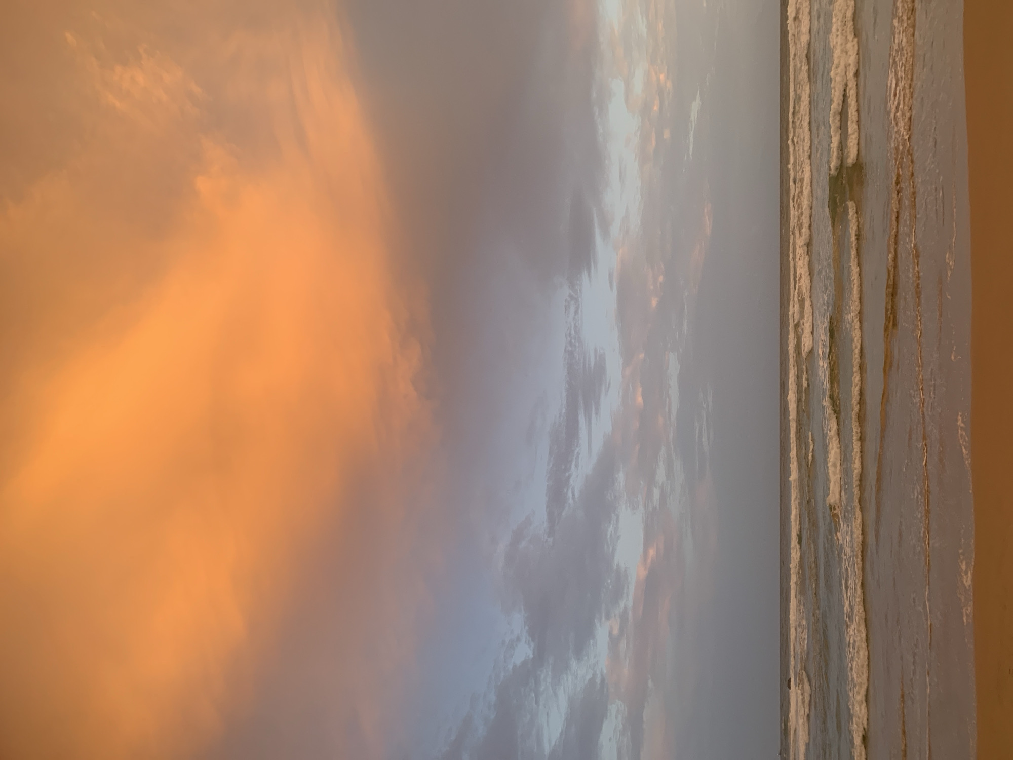 A vertical, medium shot captures a beach scene in Kathaluwa, Sri Lanka, during either sunrise or sunset. The upper portion of the image is dominated by a sky filled with clouds exhibiting a gradient of colors, ranging from vibrant orange at the top to softer pinks and grays as they descend. The lower half of the frame shows the ocean with gentle waves rolling onto a sandy beach. The water appears calm, with white foam cresting the waves. The sand itself is a warm, earthy tone. The scene is devoid of any people or man-made structures, emphasizing the natural beauty of the coastal environment.