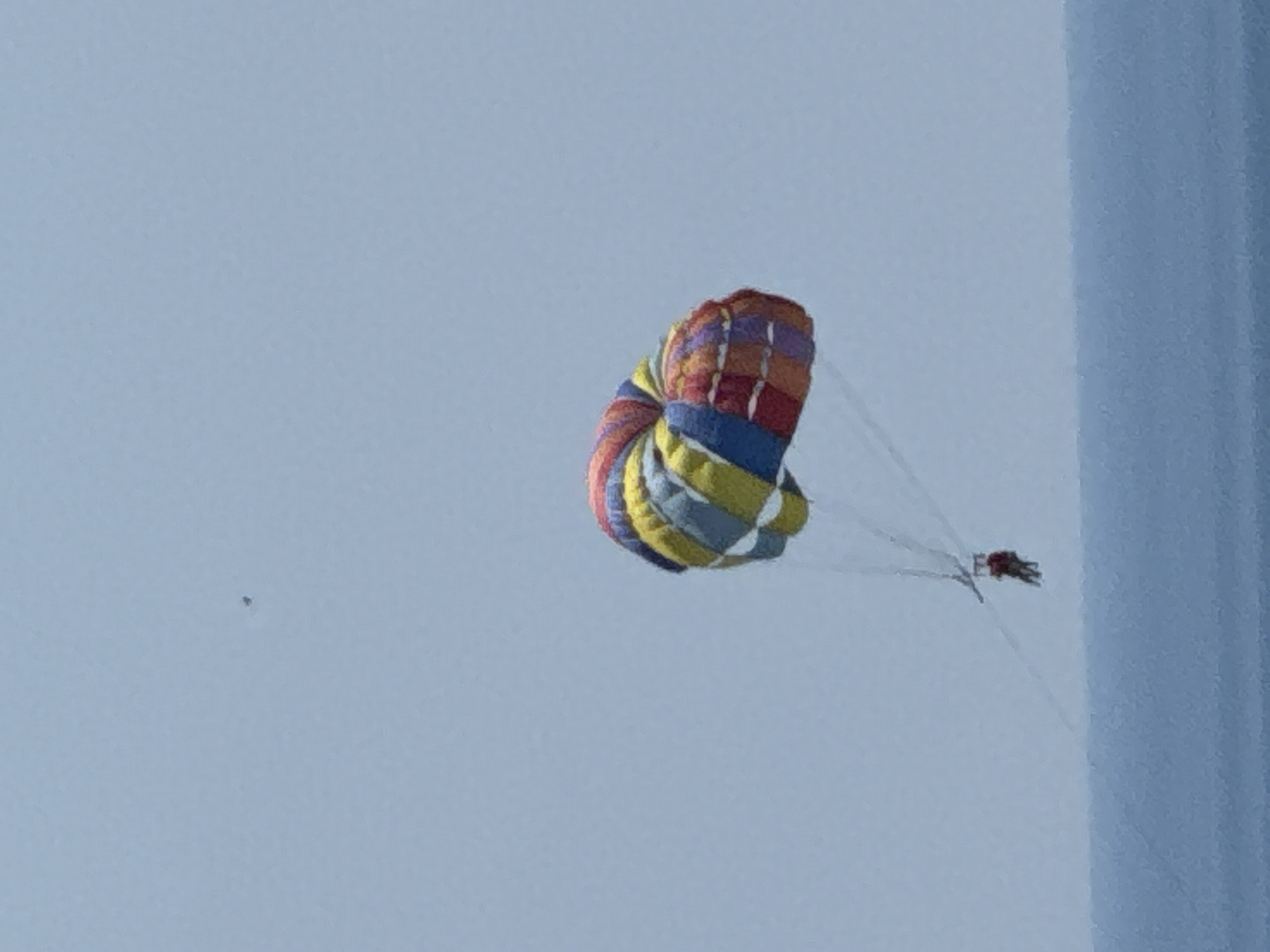 The image depicts a colorful parachute in mid-air against a pale blue sky, suggesting it is daytime and likely clear weather. The parachute is constructed from panels of various colors, including red, yellow, blue, and purple, and it appears to be in the process of being deployed or is being used for parasailing. There are visible lines extending from the parachute, indicating it is attached to something, likely a boat or a person. To the right of the parachute, a thin, vertical, light blue structure is visible, possibly a building or a large flag. In the distance, a small, dark speck is present in the sky, which could be a bird or another object. The overall scene evokes a sense of leisure and outdoor activity. The location context of Boreti, Montenegro, suggests this might be a tourist area, commonly associated with water sports like parasailing. No visible text is present in the image.