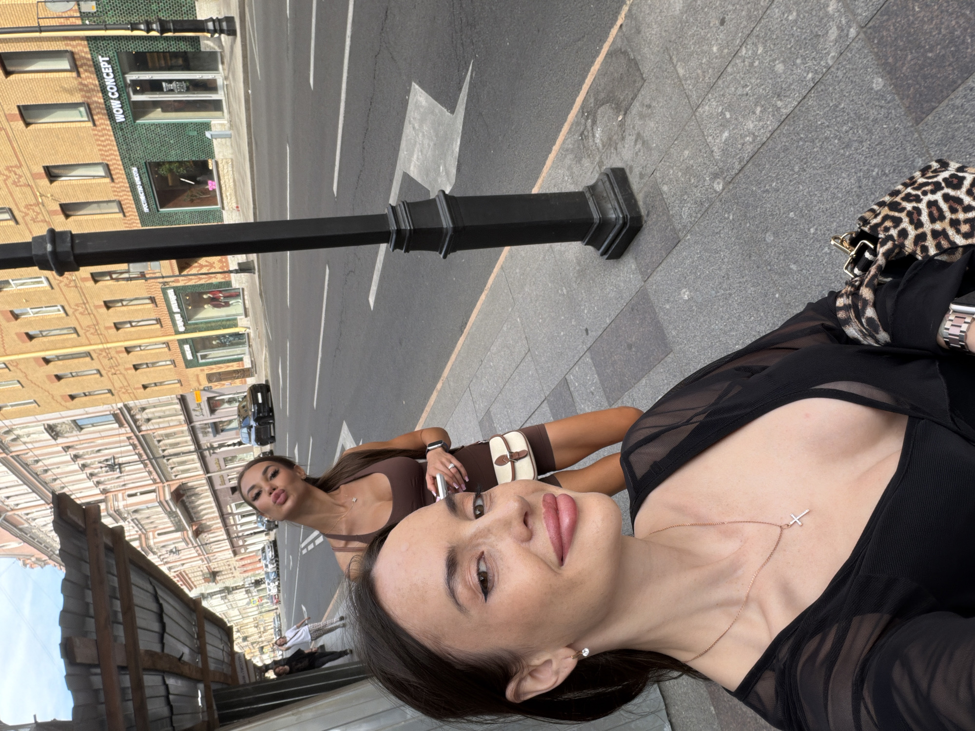 The image depicts an urban street scene in Saint Petersburg, Russia, specifically on Nevsky Prospekt, with the camera held by a woman in the foreground taking a selfie.
In the foreground, an adult female with dark hair, freckles, and pronounced pink lips smiles subtly. She wears a sheer black long-sleeved top over a darker garment, a rose gold chain necklace with a small cross pendant, and a single diamond stud earring in her left ear. Her left wrist bears a rose gold watch, and she carries a leopard-print bag in her right hand.
Behind her and slightly to the right, another adult female with long dark hair pulled back into a ponytail makes a "kissing" face at the camera. She wears a form-fitting brown sleeveless top and matching brown shorts, a thin gold necklace, a ring on her left ring finger, and a watch on her left wrist. She holds a silver object in her right hand and a small, beige and white crossbody bag with a flap closure.
To the far left in the background, two additional individuals walk on the sidewalk, moving away from the camera. The street is lined with multi-story historic buildings; those on the immediate left have a metallic corrugated awning above the sidewalk.
Across the street, a prominent yellow brick building features multiple windows. At ground level, there are retail establishments. One shop, with a distinctive green tiled facade, has Cyrillic text visible, including "КРАСОТЫ" (beauty). Adjacent to it, another shop clearly displays "WOW CONCEPT" in English.
A black, ornate street lamp post stands upright on the right side of the image, separating the grey paved sidewalk from the asphalt road. The road features white dashed lane markings and a large white arrow painted on the surface, indicating a left turn. An orange line marks the edge of the sidewalk where it meets the road. The scene is illuminated by natural daylight, suggesting late morning or afternoon.