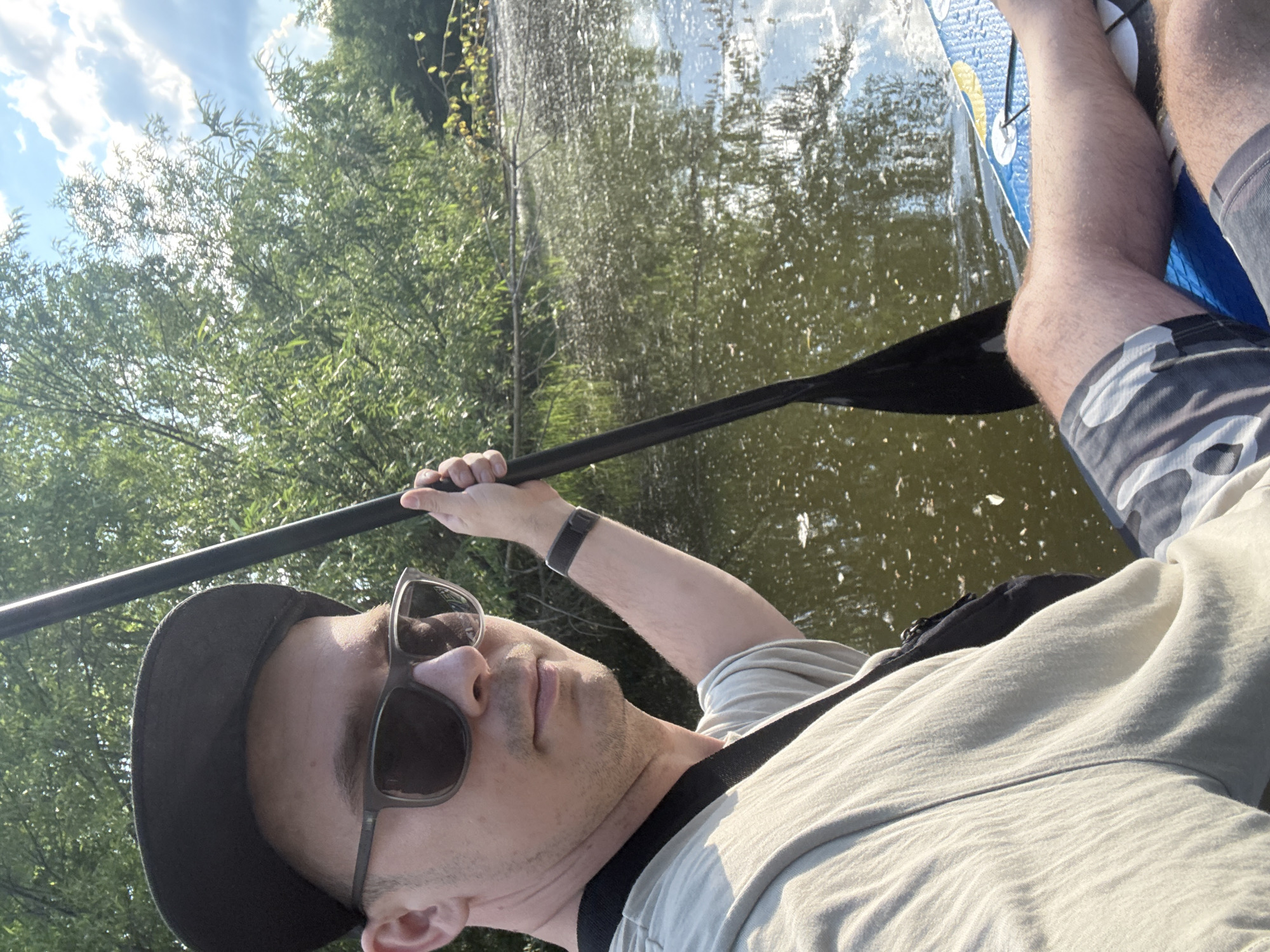 A selfie of a man on a paddleboard in a body of water, likely a river or lake, in Kondratovo, Russia. The man is wearing sunglasses, a black baseball cap, and a light-colored t-shirt. He is holding a paddle and is positioned in the foreground of the image. The background consists of dense green trees and a cloudy sky. The water appears murky with some debris floating on the surface. The paddleboard is visible in the lower right portion of the frame.