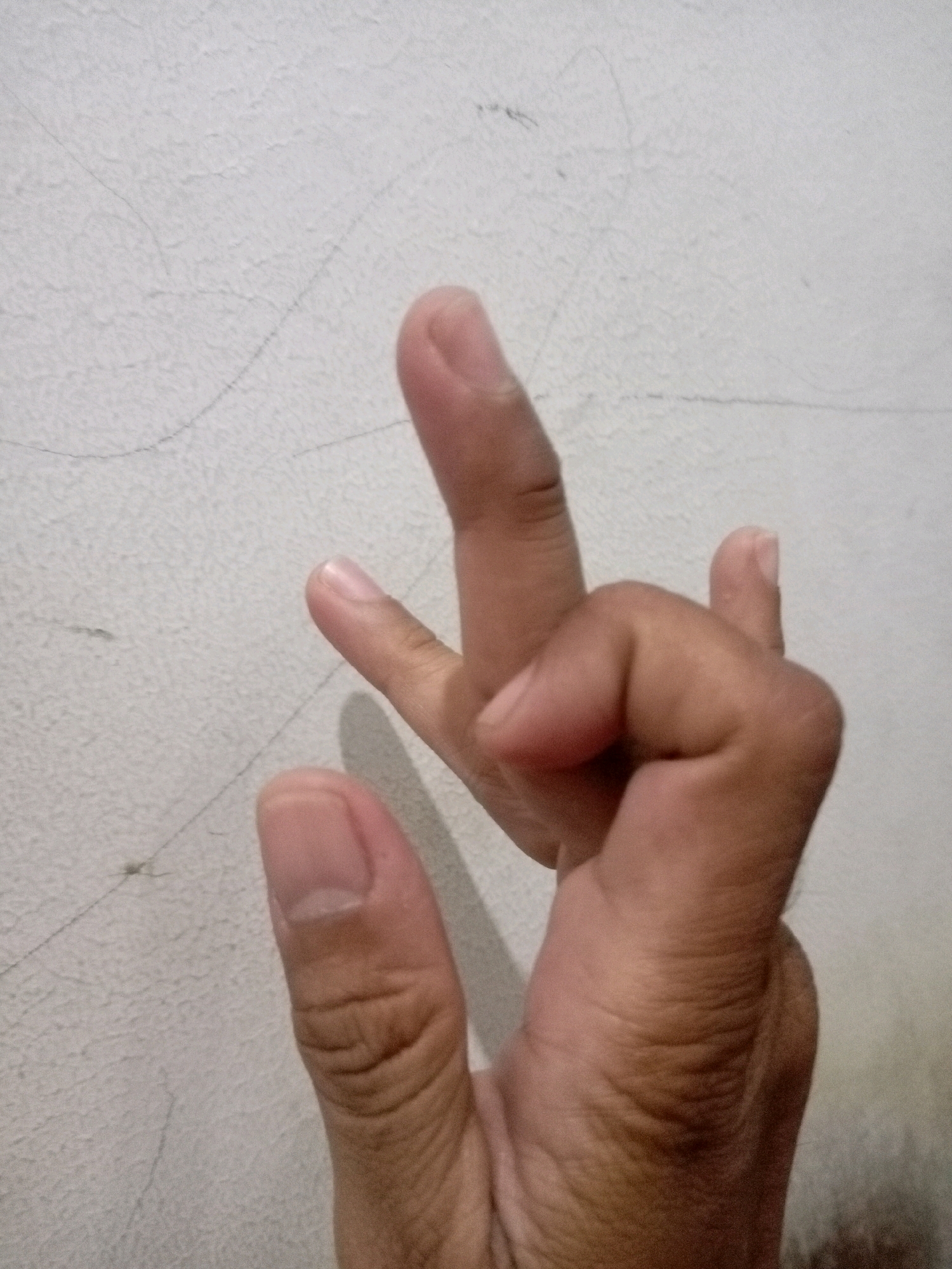 The image shows a close-up of a person's hands against a textured white wall. The hands are positioned in a way that suggests a gesture or pose. One hand has fingers bent, with the index finger extended upwards and the middle finger bent downwards, almost touching the palm. The other hand, partially visible, has a thumb extended. There are faint dark lines on the wall, possibly indicating cracks or marks. The lighting suggests an indoor environment. The location is identified as Batam, Indonesia.