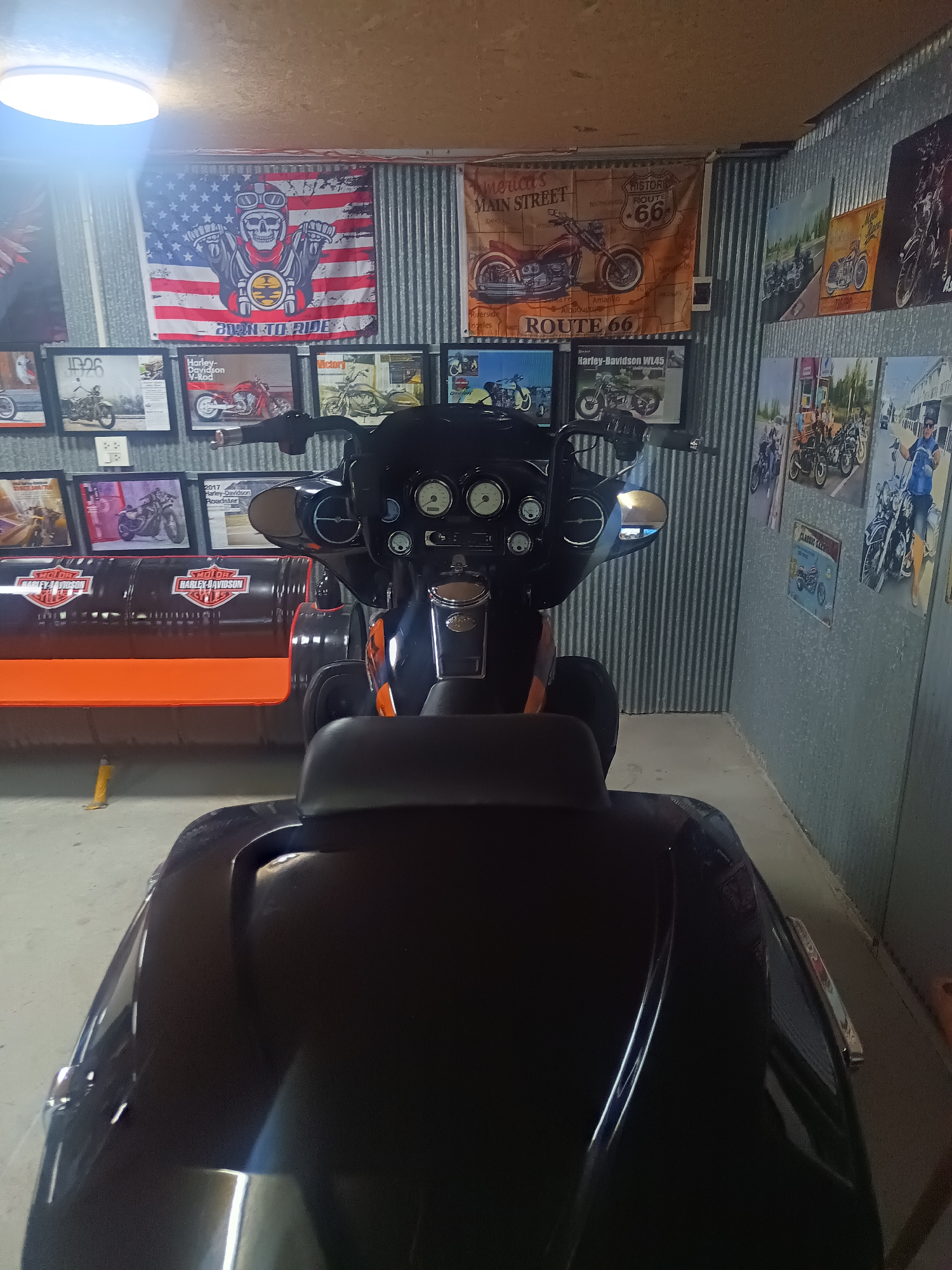 The image captures an indoor space decorated with motorcycle memorabilia. The primary focus is a black motorcycle, positioned centrally and taking up much of the frame. The seat and rear storage unit are clearly visible, obscuring some of the background elements.
Behind the motorcycle, the wall is covered with a combination of metal sheeting and decorative items related to motorcycles. Framed motorcycle images, posters, and flags are arranged along the wall. Notably, there is a flag depicting a skull wearing a motorcycle helmet with the words "BORN TO RIDE" printed on it, along with a "ROUTE 66" flag.
To the left, there is a makeshift bench constructed from oil barrels, painted black with orange seating. The lighting in the room is provided by an overhead ceiling light.
The location is identified as Surat Thani City Municipality, Thailand, based on the prompt.