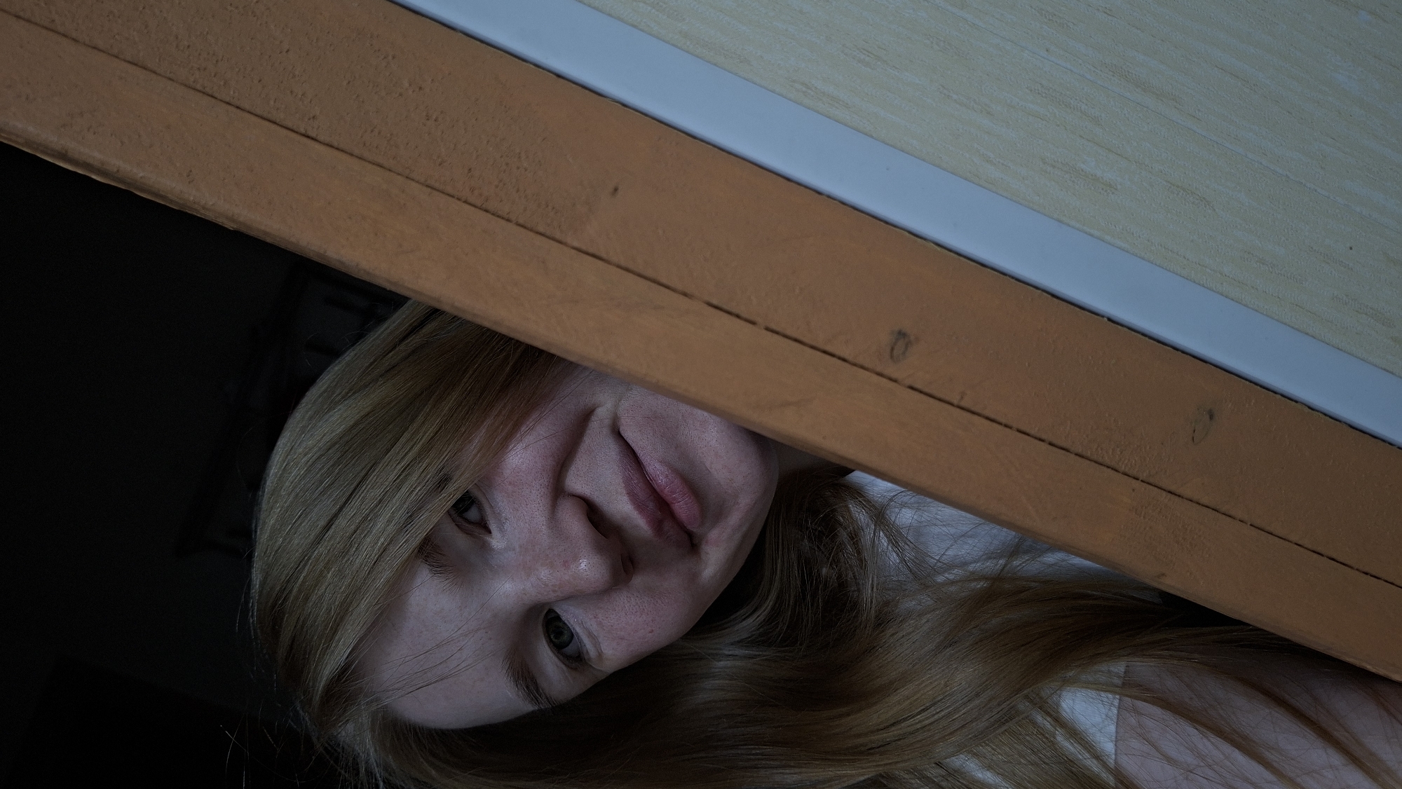 The image shows a close-up, low-angle selfie of a young woman with long, light brown hair, peeking from behind an orange-brown wooden beam. She has fair skin with freckles and is looking directly at the camera with a slight smile. Her right eye is visible, and the left side of her face is obscured by her hair and the beam. She is wearing a white top. The background is dark, suggesting an indoor setting, potentially with low lighting. There are no identifiable landmarks or elements that would suggest a specific location within the city of Большой Букор, Russia.