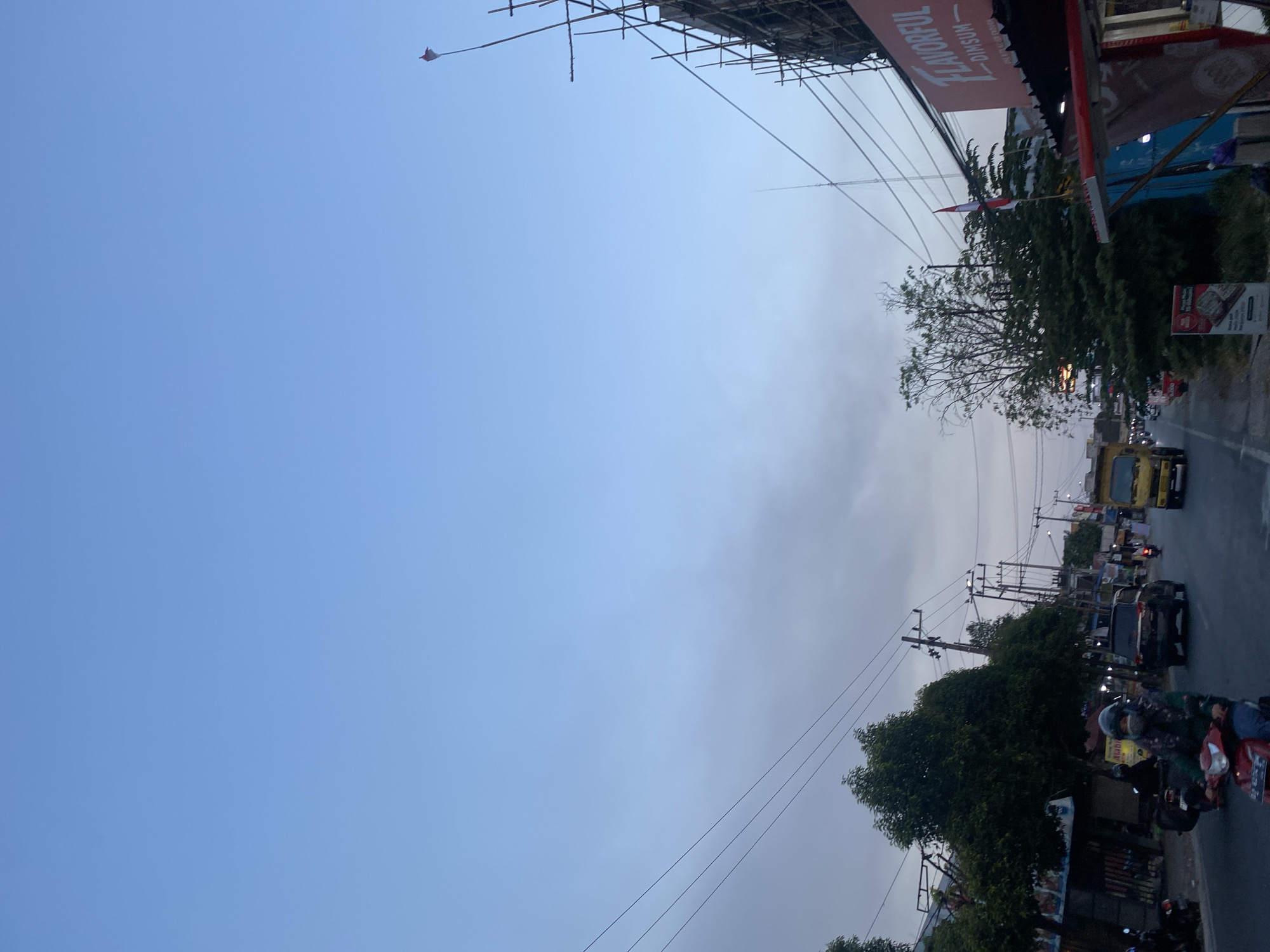 In Banjarbaru, Indonesia, a road is captured at dusk, with a clear, pale blue sky transitioning into dusk hues. A red and white Indonesian flag flutters atop scaffolding on the right side of the frame, near a sign that reads "FLAVORFUL DIMSUM." The road is lined with trees and utility poles. Traffic moves along the asphalt road, including a red motorcycle with a masked rider in the foreground, a black SUV, and a yellow truck. The scene appears to be a commercial area with various signs and awnings visible.