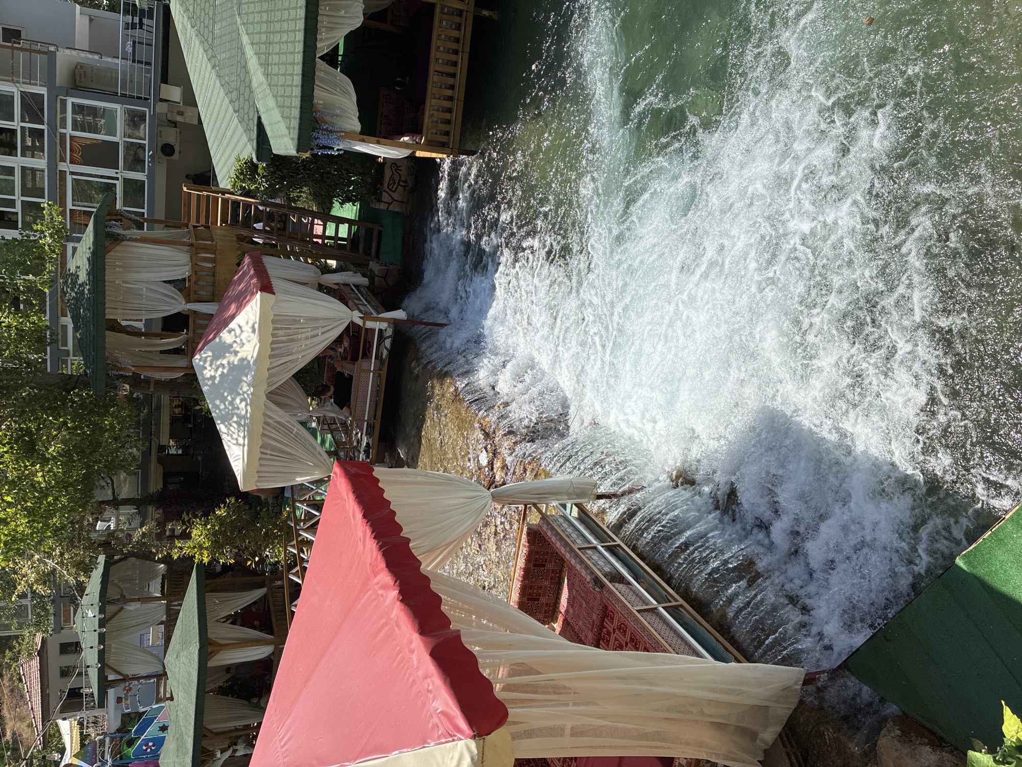 The image depicts a picturesque outdoor dining area situated over a flowing river with waterfalls. Several open-air gazebos, furnished with cushioned seating and decorative curtains, are strategically placed along the riverbank and over the water. Some gazebos feature red and white striped canopies, while others have green roofs. The water is clear and turbulent, with noticeable cascades. Trees and buildings are visible in the background. One building appears to be a multi-story structure with numerous windows. A wooden staircase leads up to one of the elevated gazebos. A few individuals can be seen seated in the gazebos. The setting suggests a resort or restaurant in a natural, water-centric environment. This location is likely Dimçayı River valley, a popular tourist spot near Alanya, Turkey, known for its riverside restaurants and recreational facilities.
