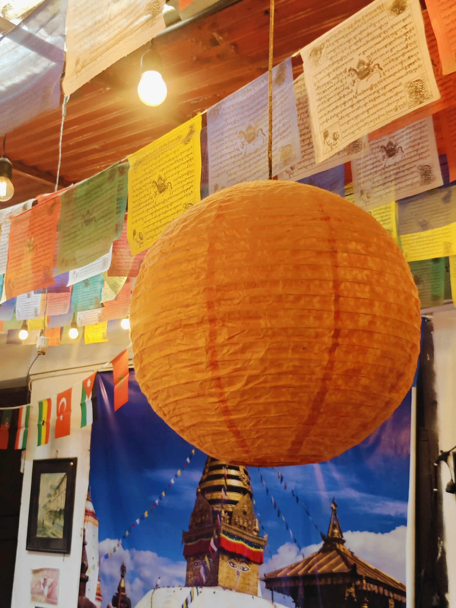 The image captures an indoor scene, likely a cafe or shop, decorated with prayer flags and featuring a large, orange paper lantern as the central subject. The prayer flags, printed with text and images, are strung across the ceiling, some appearing weathered. Several exposed light bulbs hang from the ceiling, illuminating the space. In the background, a large mural depicts a prominent Buddhist stupa, characteristic of Kathmandu, Nepal, under a bright blue sky with clouds. Small national flags are also visible, strung along a wall. The overall ambiance suggests a cultural or tourist-oriented establishment in Kathmandu.