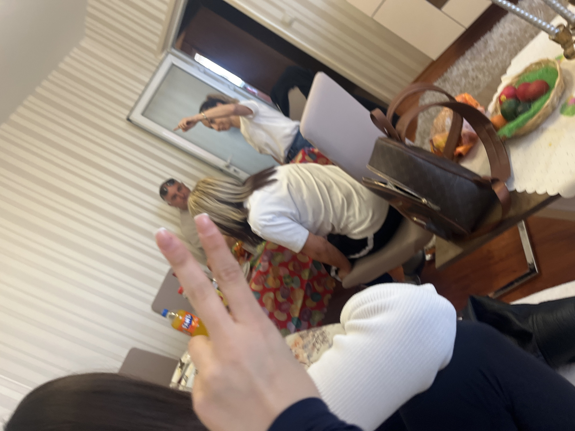 The image depicts an indoor scene with several individuals. A person in the foreground, wearing a white hat and a dark blue garment, is making a peace sign with their fingers. Behind them, two people are visible: one is pointing, and the other is wearing a white shirt. In the background, another person is partially visible. There is a table with a purse and a basket with fruits. A bottle of Fanta is placed on a surface. The location is potentially Varna, Bulgaria.