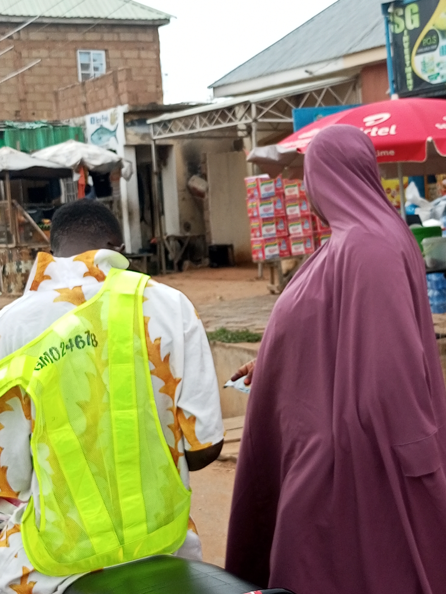 The image depicts a street scene in Tempure, Nigeria. Two individuals are partially visible. On the left, a person wearing a white shirt with an orange pattern and a bright yellow safety vest with the text "GMD24678" is seen from the back. On the right, a person is wearing a purple hijab and cloak, seen from the side and slightly from behind. In the background, there are buildings with brick and corrugated metal construction, market stalls with umbrellas, and stacked goods, including what appears to be cases of bottled water and packaged food items. A red umbrella with the "Artel" logo is prominent. The environment appears to be an outdoor market or a busy street with commercial activity.