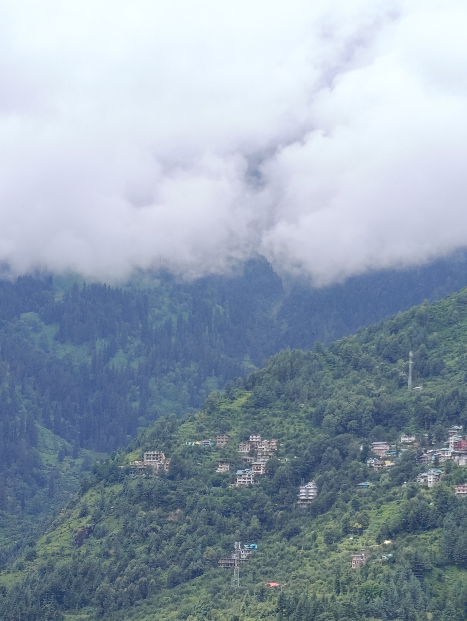 A full shot captures a mountainous landscape with a thick forest covering the slopes. The sky is overcast with clouds that partially obscure the mountain peaks. Several buildings, appearing to be houses or small hotels, are scattered across the mountainside, blending into the natural environment. Some of these structures are clustered together, forming small hamlets, while others are more isolated. There is a prominent communication tower visible on one of the slopes, indicating some level of infrastructure within this remote setting. The overall impression is of a serene, yet inhabited, natural landscape. This scene is located in the city of Kullu, India.