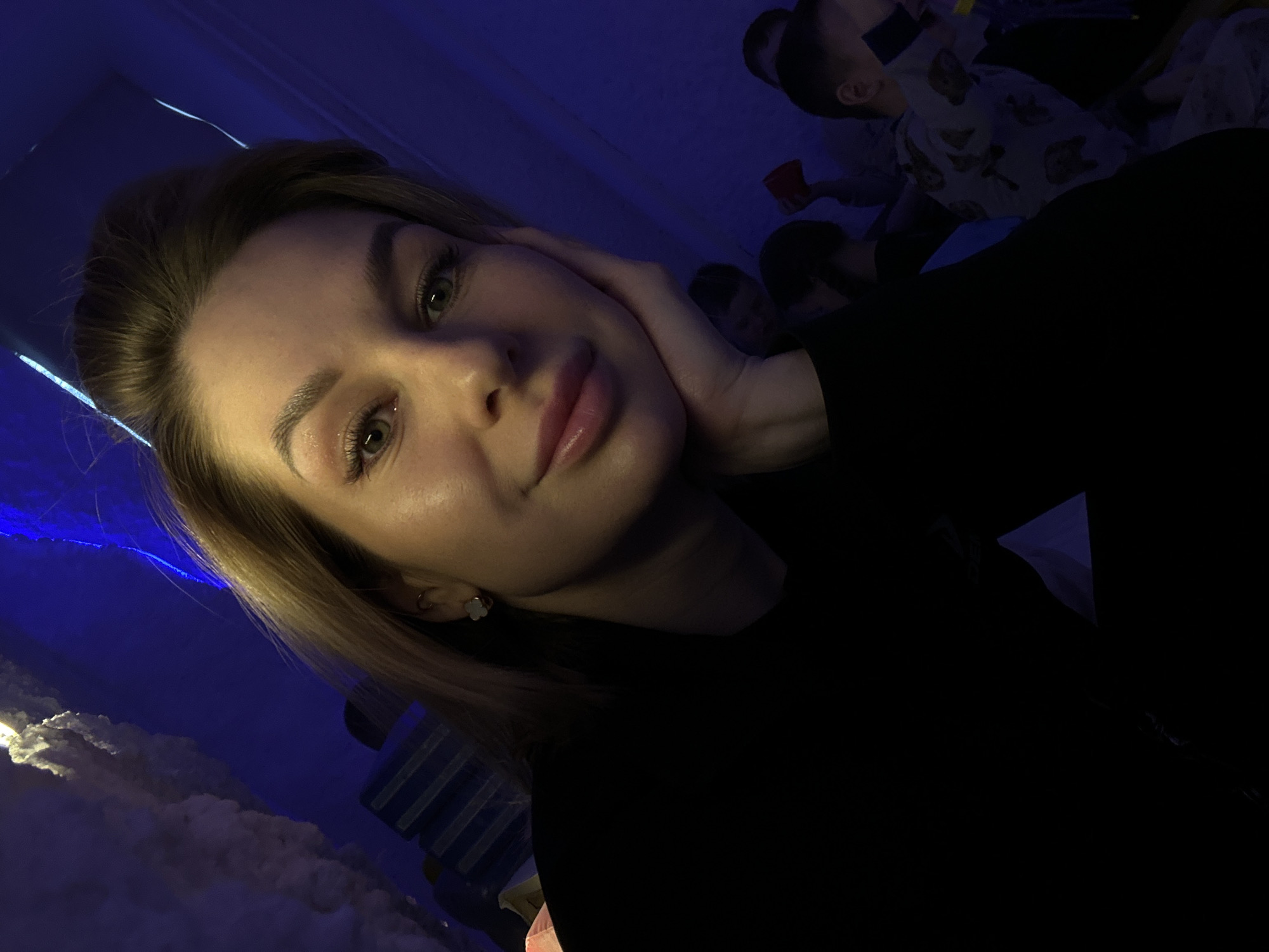 A woman with light brown hair and green eyes is in the foreground of the photo, holding her face with her hand. She's wearing a black top. Behind her are several people, including a child in pajamas. The environment appears to be indoors, possibly a playroom, with blue lighting and decorative cloud-like elements. There is no verifiable information to pinpoint the exact location in Chaikovsky, Russia.
