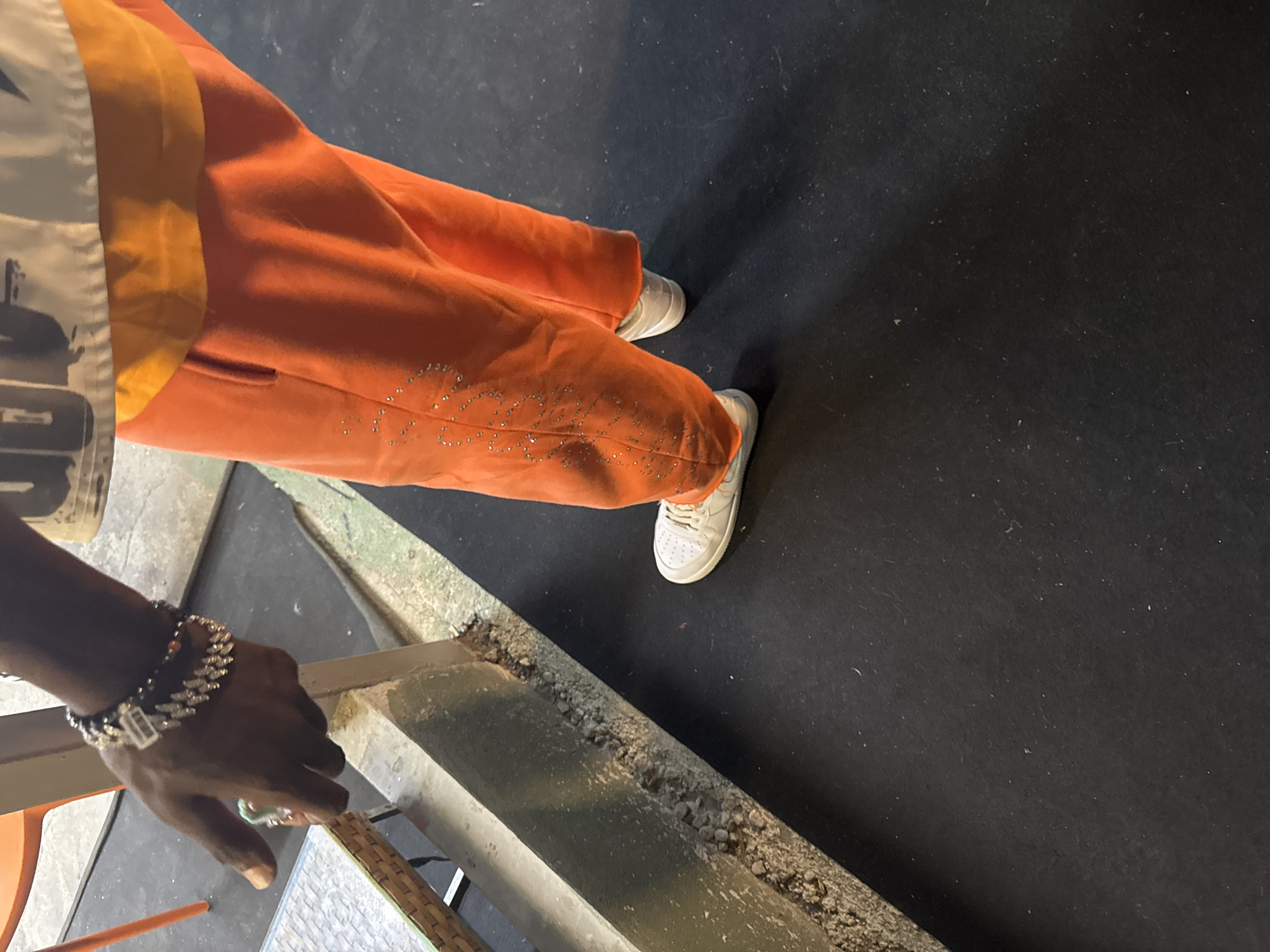 This is a close-up, low-angle, outdoor shot of a person's legs and arm. The subject is wearing bright orange sweatpants adorned with rhinestones, white sneakers, and a bracelet on their left wrist. They are standing on a dark, textured surface, possibly a mat or pavement. To the left, a portion of a table with a woven surface is visible. A dark, textured vertical beam runs along the left side of the frame, with rough, broken concrete at its base. The lighting suggests it is daytime, with shadows cast on the ground. The location is not identifiable as Aksu, Turkey based on the visual information provided.