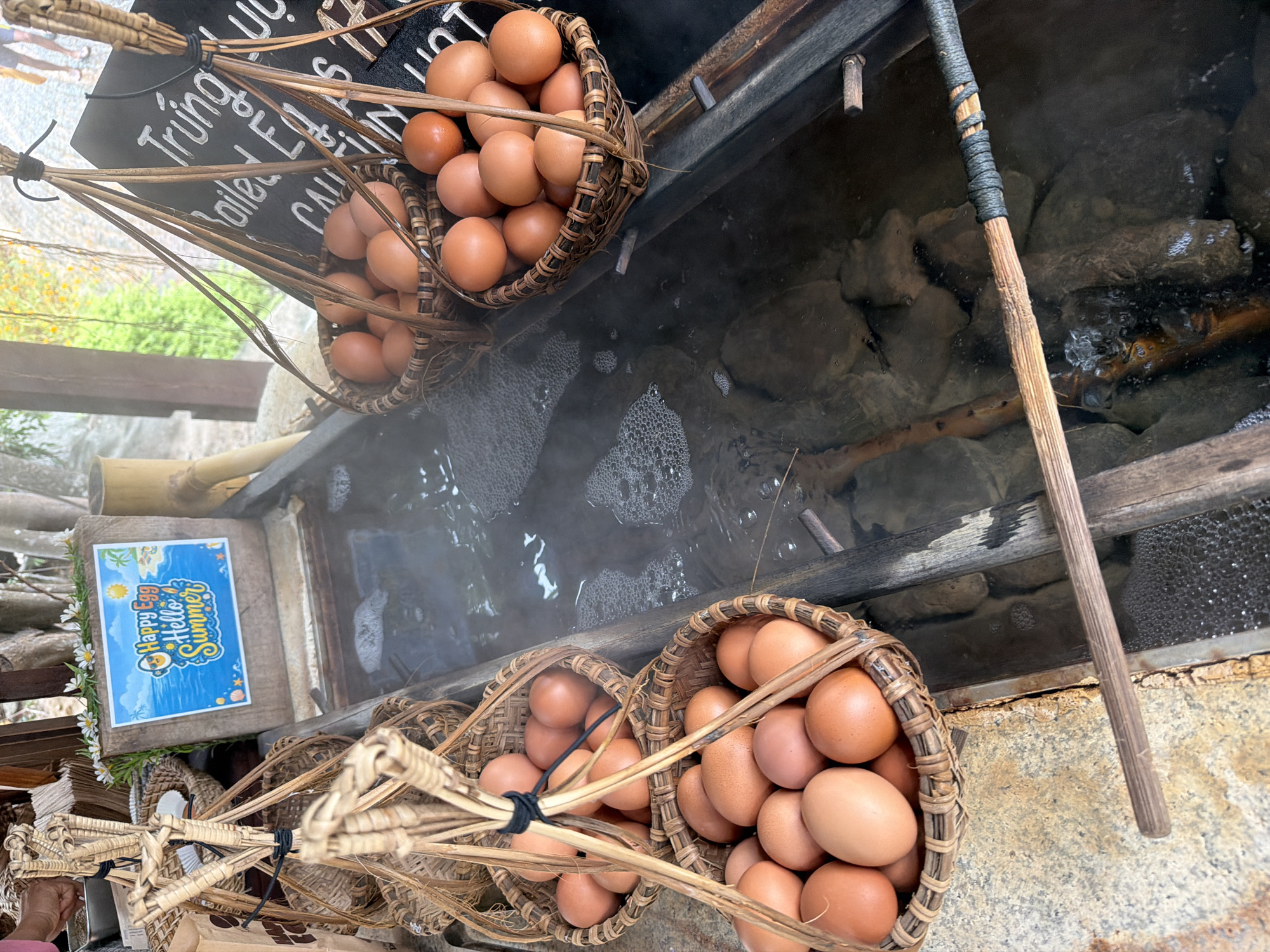 A rustic setup shows several woven baskets overflowing with brown eggs being lowered into a hot spring in Nha Trang, Vietnam. Steam rises from the mineral-rich water, indicating it is hot enough to cook the eggs. A wooden sign with the text "Happy Egg Hello Summer" is visible in the background, adding a festive touch to the scene. The surrounding environment appears natural, with rocks and foliage suggesting an outdoor setting. The process appears to be a traditional method of cooking eggs using the geothermal heat of the spring.