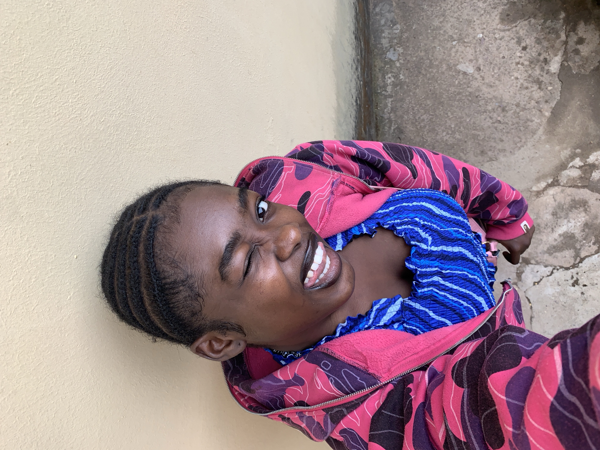 A woman is smiling and winking at the camera. She is wearing a pink and purple patterned jacket over a blue striped top. She is leaning against a beige wall. The ground is gray concrete. The location is Rafin Par, Nigeria.