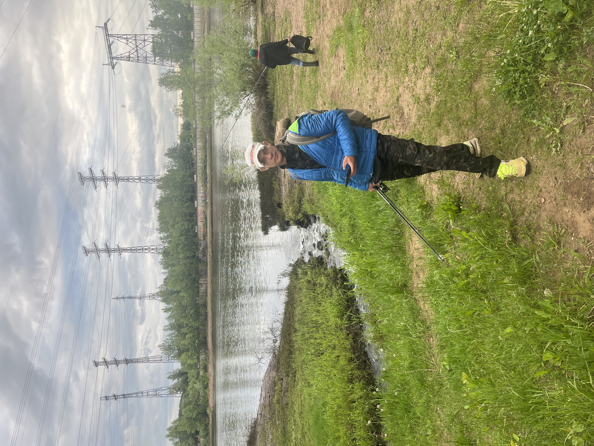 This image depicts an outdoor scene in the city of Chaikovsky, Russia, on an overcast day. The primary subject is an adult male standing in the left-center foreground, facing slightly towards the viewer. He wears a white baseball cap with red accents, a bright blue puffer jacket, a dark patterned shirt, dark camouflage-patterned pants, and light-colored athletic shoes (one yellow/lime green). He carries a grey/olive backpack with a visible green top accent on his right shoulder. In his left hand, he holds a fishing rod angled downwards, and in his right hand, he holds a dark smartphone. His face shows stubble.
The environment immediately surrounding him consists of a grassy bank with patches of bare earth and white dandelion flowers. To his left, a small stream or ditch with flowing water cascades over rocks before merging into a larger, calm body of water.
In the mid-ground, a second adult male is observed from behind, walking towards the right along a dirt path bordering the water. He wears a dark jacket, blue jeans, dark rubber boots, and a green baseball cap. He carries a dark bag in his left hand and appears to have a fishing rod extended towards the water.
The large body of water occupies the middle ground, reflecting the grey, cloudy sky. Across the water, a dense line of green deciduous trees runs horizontally. Behind the trees, a concrete retaining wall with some faded graffiti is visible. Dominating the distant background are five prominent metal lattice electricity transmission towers, carrying multiple power lines across the landscape under the heavily overcast sky.