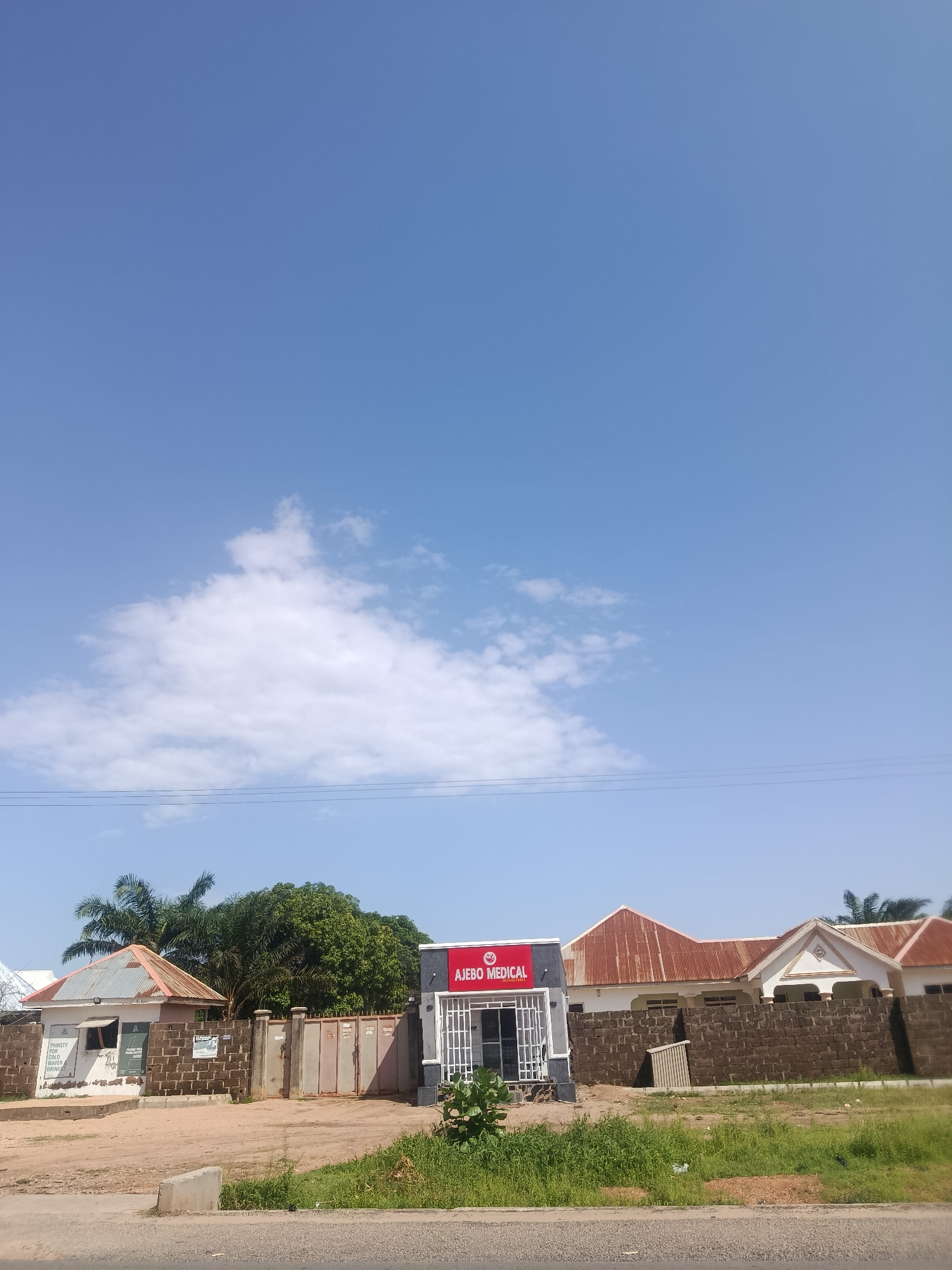 The image depicts a street view of a commercial establishment in Jalingo, Nigeria. On the left, a small white building with a corrugated iron roof has a sign that reads "THIRSTY FOR ICE COLD WATER DRINKING HERE". Next to it is a gate made of metal bars. To the right, a modern-looking building with a red sign displaying "AJĘBO MEDICAL" is visible. This medical facility appears to be part of a larger complex, with adjacent buildings featuring rusted corrugated iron roofs and dark brick walls. The scene is set against a backdrop of lush green trees and a clear blue sky with scattered white clouds. The ground in front of the buildings is dusty and unpaved.