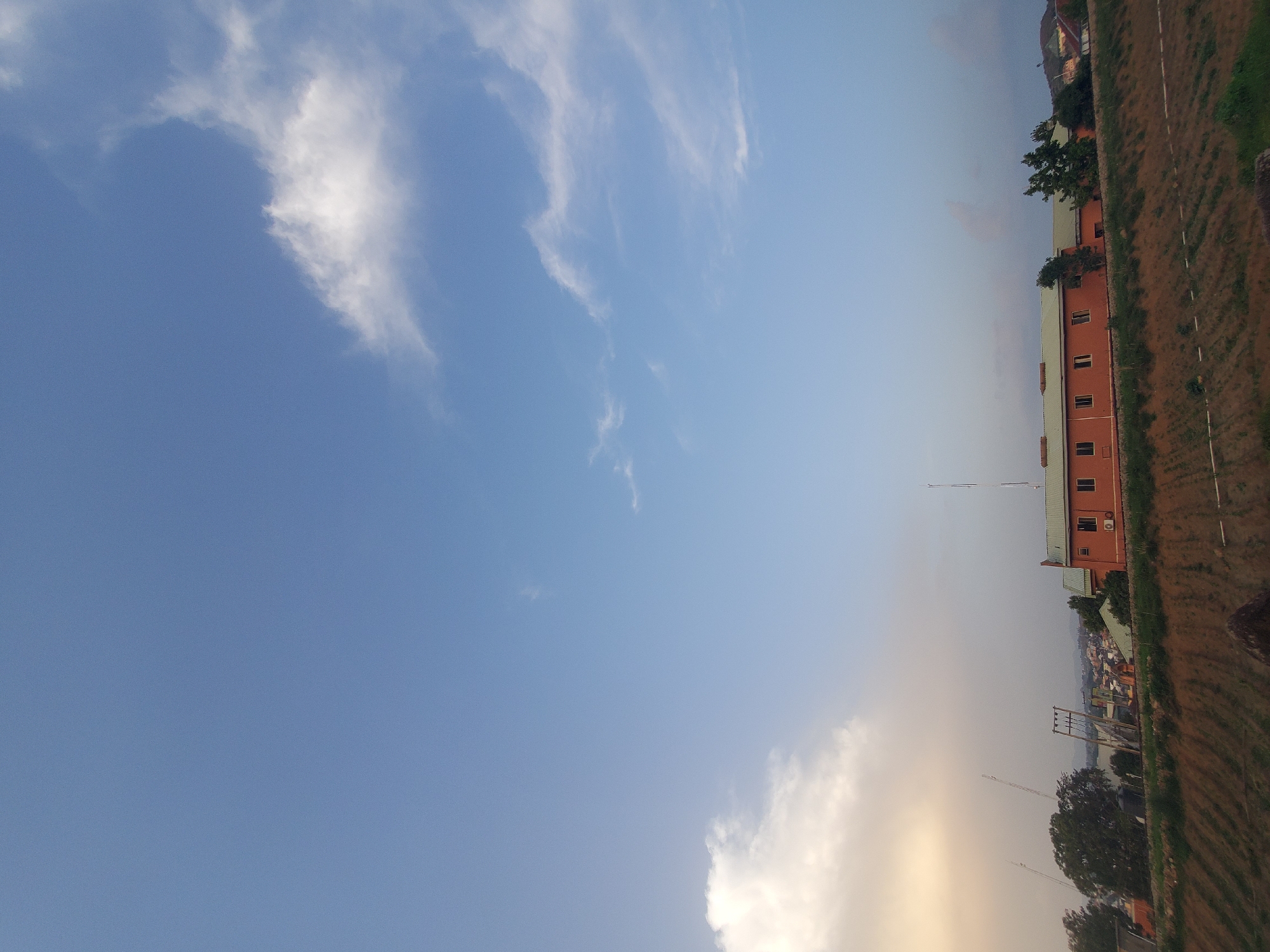 This image captures a clear blue sky with scattered white clouds over a distant cityscape in Jos, Nigeria. In the foreground, a dry, tilled field stretches across the bottom of the frame. A low, orange building with a green roof, featuring multiple windows and an air conditioning unit, is situated in the middle ground. Several tall, slender structures, possibly communication towers or light poles, are visible behind the building and to its left. Trees and other buildings are scattered throughout the landscape, suggesting an urban or semi-urban environment. The overall impression is one of a daytime scene with a wide expanse of sky dominating the upper portion of the image.