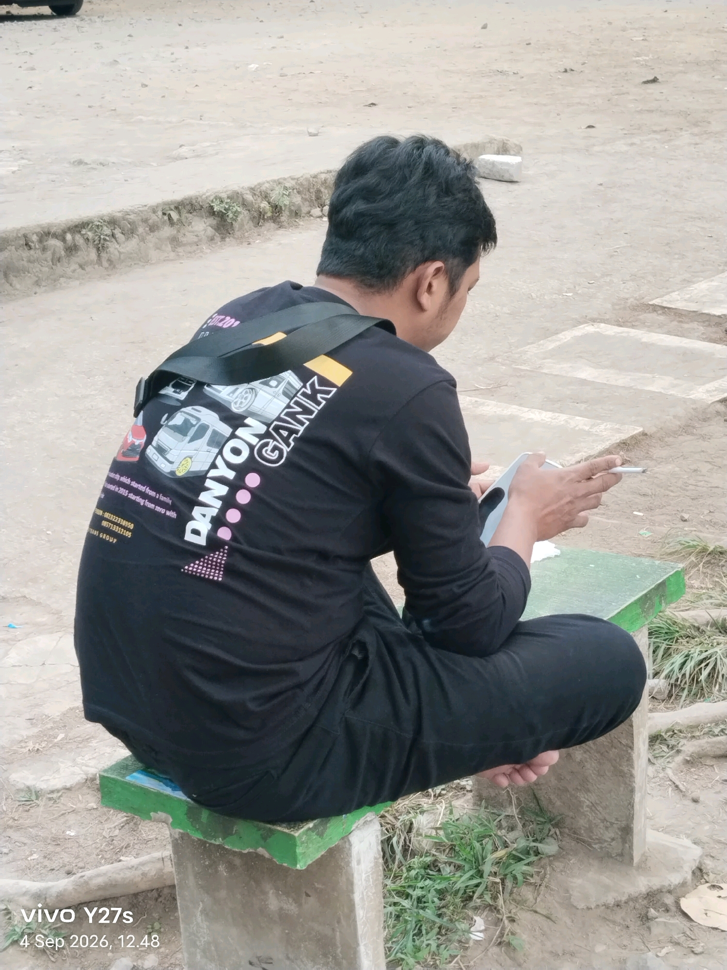 A person is sitting cross-legged on a concrete bench with a green top in Maron, Indonesia. The person is wearing a black long-sleeved shirt with a graphic design on the back, a black bag slung over their shoulder, and black pants. The person is holding a smartphone in their left hand and a cigarette in their right hand. The ground around the bench is dirt and gravel with some patches of grass. The image has a timestamp in the bottom left corner indicating it was taken on September 4, 2026, at 12:48 PM.