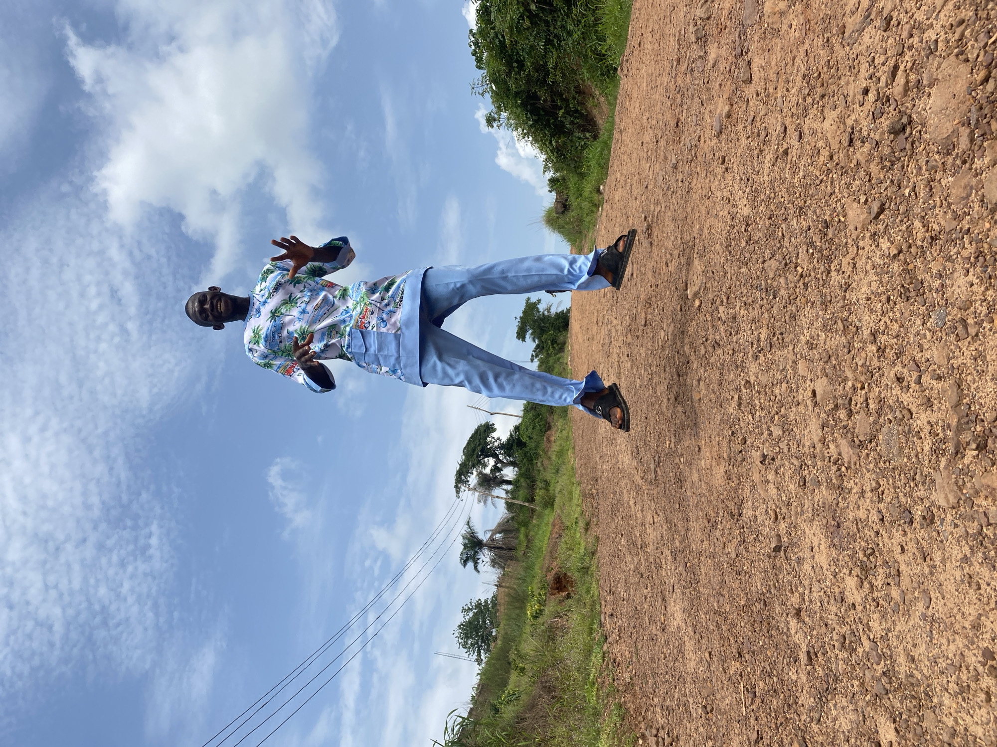 An individual wearing a patterned, light blue and white shirt, light blue pants, and dark sandals stands on a dirt road. The sky is visible with blue and white clouds. Trees and shrubs line the side of the road. The location appears to be in a rural area of Ufu Esioni, Nigeria. Power lines run along the left side of the scene.