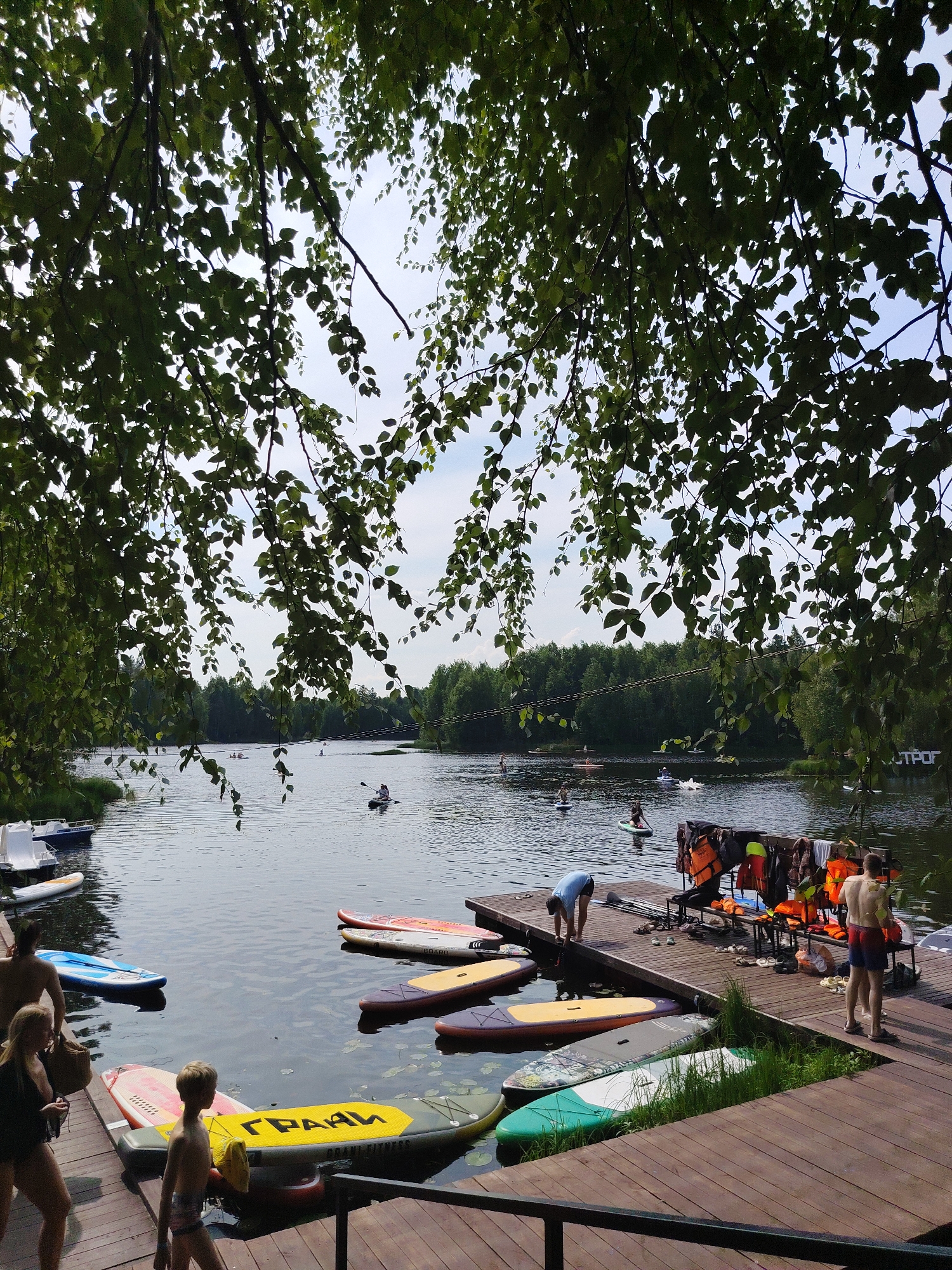 This image depicts a recreational waterfront scene at a lake or river, likely in or near Vylytydor, Russia, given the Cyrillic text on a paddleboard that reads "ГРААН" (GRAAN). In the foreground, a wooden dock is visible, with several paddleboards and a young boy standing on it. Further back, a woman and a man are walking on the dock. The water is dotted with several people on paddleboards and kayaks, engaged in various activities. A rack with life jackets is also present on the dock. The background features a dense line of green trees under a cloudy sky. The overall atmosphere suggests a summer day at a watersports facility.