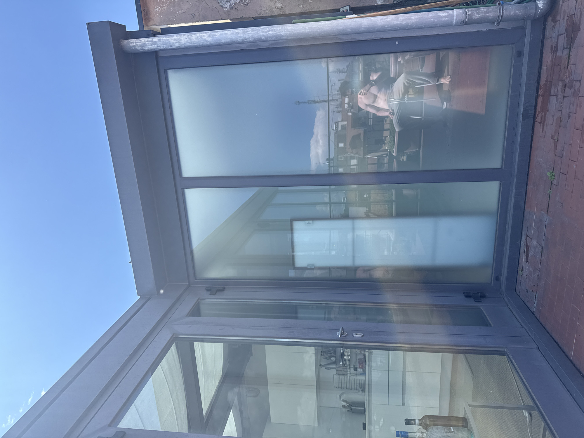 A low-angle shot showcases the exterior of a modern architectural structure, possibly a penthouse or an addition to an existing building, located on a rooftop in Milan, Italy. The structure is defined by a clean, contemporary design featuring floor-to-ceiling glass panels framed by sleek, gray-toned metal supports. The glass reflects the surrounding urban landscape, including other buildings and a clear blue sky. In the reflection, there is a shirtless man working out or posing with his back to the viewer. The exterior roof is covered in bricks and the structure has a large gutter system in place. There are various cleaning supplies inside the apartment as well.