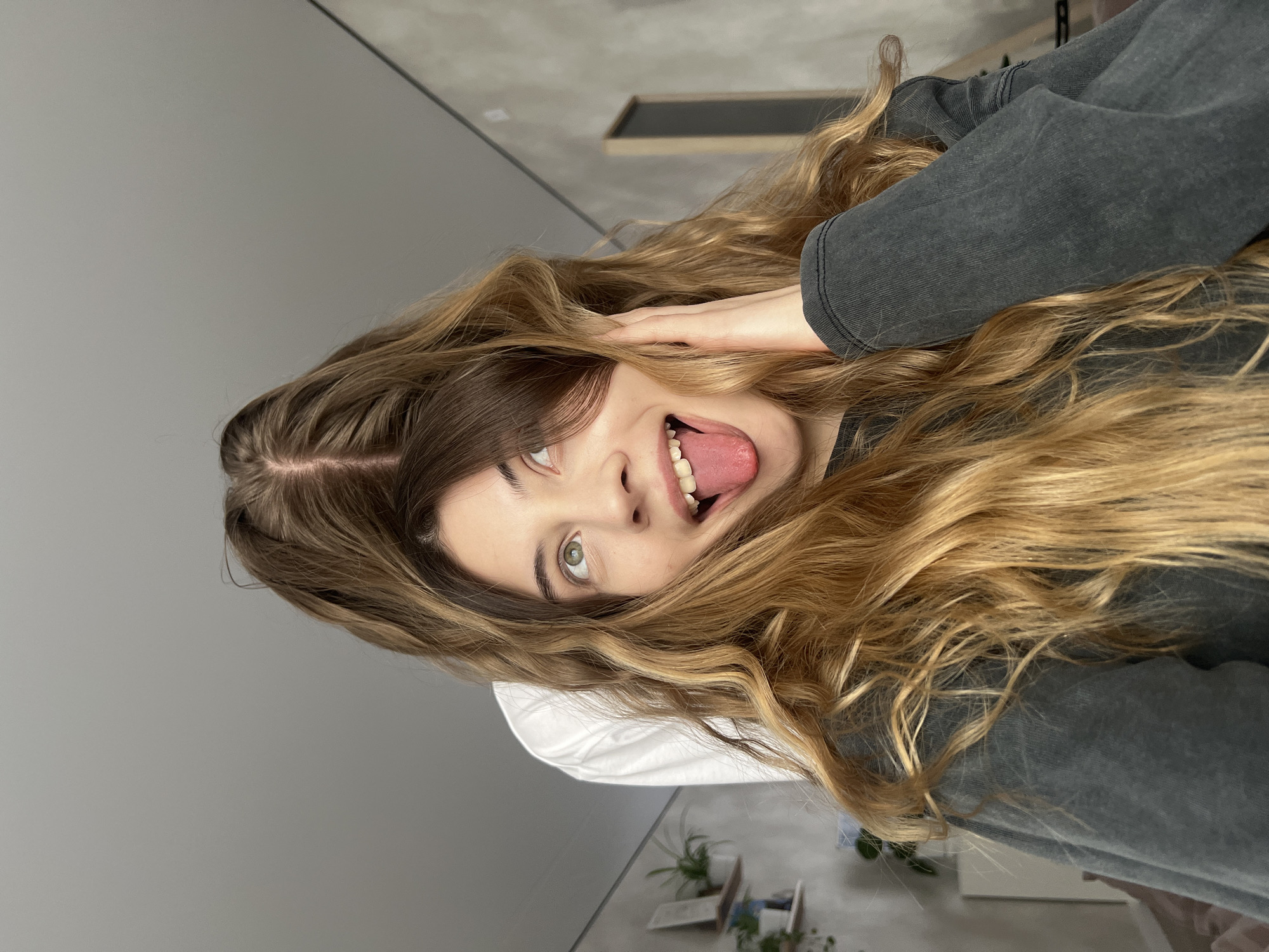 A person with long, wavy, ombre hair is lying on a white pillow. She is sticking out her tongue and touching the side of her head. She is wearing a dark gray sweatshirt. The background consists of a gray wall, a small framed picture, and a potted plant. The location is Unknown, Unknown.