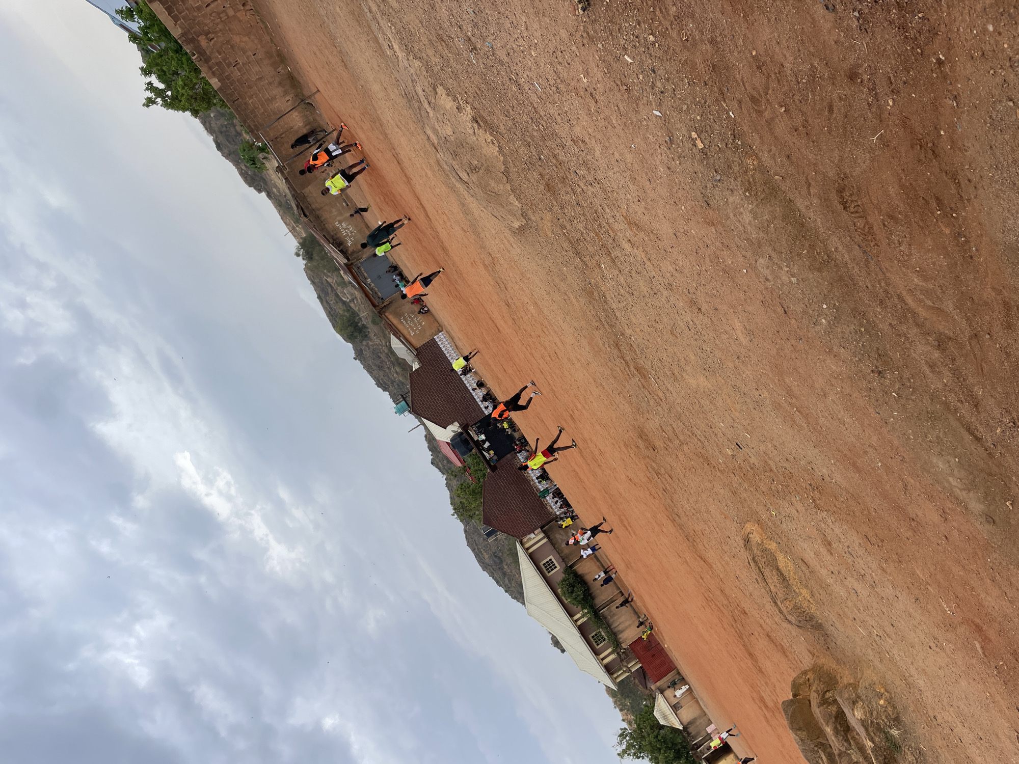The image captures an outdoor scene in Jos, Nigeria, characterized by a wide expanse of reddish-brown terrain. A building with multiple sections and rooflines is positioned along the edge of the terrain. A group of individuals is arranged in a line adjacent to the building. Some of them are wearing bright athletic clothing, while others have darker clothing. They appear to be engaged in some form of physical activity. The sky is overcast with a multitude of gray clouds. A hill rises in the background.