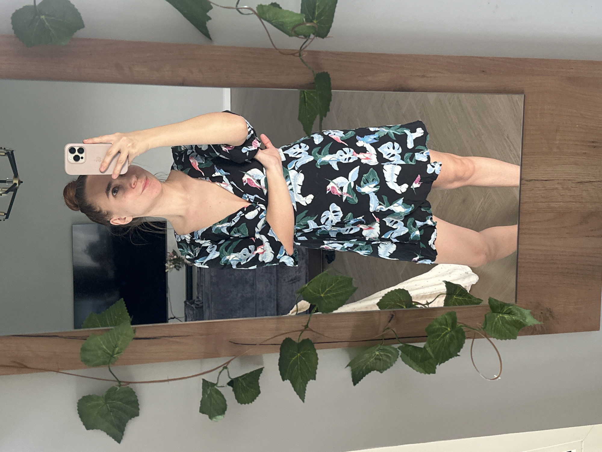 A woman is taking a selfie in a mirror. The mirror has a wooden frame and is decorated with artificial vines with green leaves. The woman has her hair in a bun and is wearing a black dress with a floral print. She is holding a pink smartphone in her right hand, pointed towards the mirror. In the background, there is a television, a cabinet, and a white blanket. The location is not discernable from the image.
