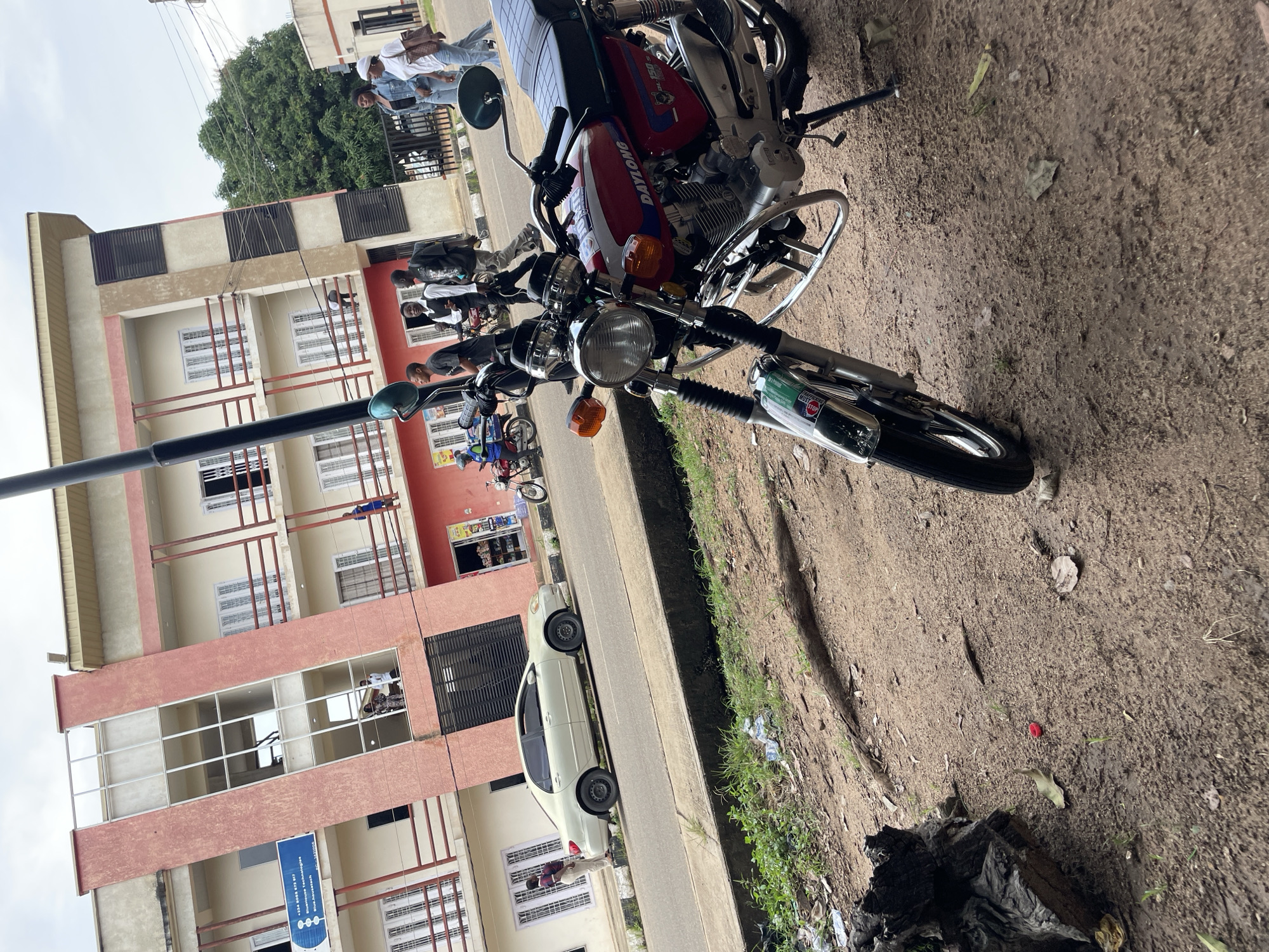 The image depicts a street scene in Jos, Nigeria, likely during daylight hours given the bright illumination. The foreground is dominated by a motorcycle, positioned at an angle and partially obscuring the view. It's a red and white model with "DAYLONG" and "150" visible on its body. The motorcycle is parked on a dirt and gravel surface, with some scattered debris.
Behind the motorcycle, a street runs alongside a series of multi-story buildings. The buildings have a reddish-pink facade with white window frames and balconies. Several people are visible on the street and near the buildings. In the upper left, a group of people, possibly pedestrians, are walking. Closer to the buildings, several individuals are gathered, some near motorcycles.
A cream-colored sedan is parked on the street, facing away from the camera. A drain or gutter runs along the edge of the street, separating it from a patch of dry ground with some green vegetation.
Notable details include a sign on one of the buildings with text that appears to be in English, including a phone number "+234 8059 871 207". The overall impression is of an ordinary day in a bustling urban or semi-urban area. The lighting suggests it's daytime, and the general appearance of the street and buildings points to a location in Nigeria. There are no strong indicators of specific weather conditions, but the scene appears dry.