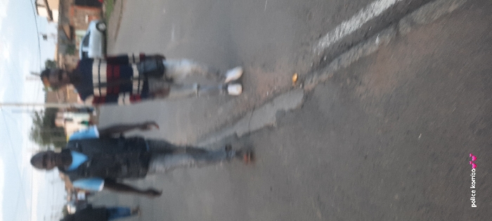 The image depicts an outdoor street scene, likely in an urban or suburban area of Nairobi, Kenya, during daylight hours. Three men are visible walking on an asphalt road, their figures slightly blurred, suggesting motion or camera shake.
In the foreground, the closest man is dressed in a dark vest worn over a light blue t-shirt, paired with light-colored trousers. Behind him, another man is seen wearing a long-sleeved shirt with horizontal stripes in dark blue/black, red, white, and light blue, along with light-colored pants and white sneakers. A third individual's lower body, clad in dark pants, is partially visible further back and to the left.
The road surface is dark asphalt, marked with a distinct white line. A small yellowish object can be seen on the road near the feet of the man in the striped shirt. In the background, a white car is parked or moving on the side of the road, and indistinct buildings or structures are visible amidst some foliage, contributing to the urban/suburban setting. The overall lighting suggests it is daytime. A watermark with the text "police kombo" and a small pink heart or flower icon is present in the lower right corner of the image.