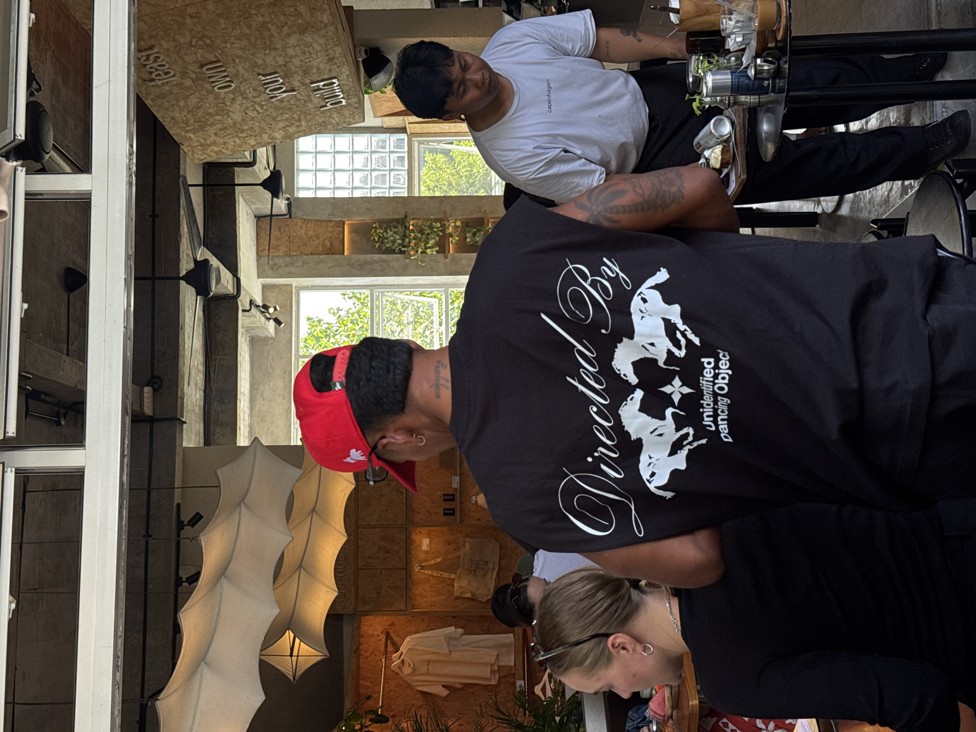 The image depicts an interior scene, possibly a cafe or retail space, with a focus on people and objects within the frame.
People: Two individuals are prominently featured. One wears a black t-shirt with the words "Directed By" and an image of white horses on the back and a red baseball cap. The other person is wearing a white t-shirt. Another individual is partly visible with blonde hair.
Objects: In the background, there are light fixtures resembling leaves, clothing items hanging on racks, and wooden panels. Above there is a sign with the words "Build Your Own" and "East". There are also clear glasses, metal cans, and other beverage containers visible.
Environment: The space is indoors with what appears to be natural light coming in through a window. The style of the space seems modern.
Location: North Kuta, Indonesia.