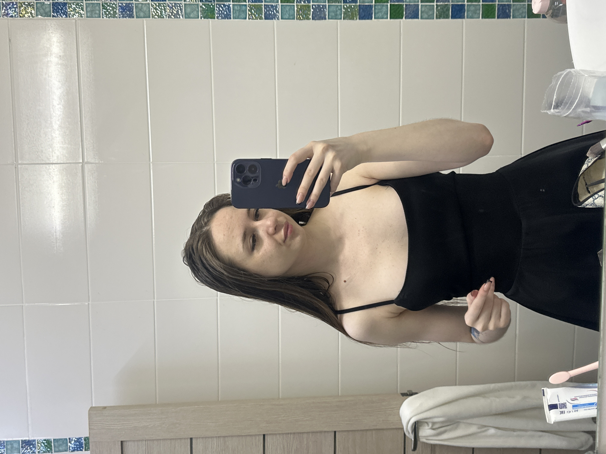 A young woman with long, dark hair is taking a mirror selfie in a bathroom. She is wearing a black tank top and a black skirt. She is holding a dark-colored smartphone in her right hand and is looking at the camera. Her left arm is bent, and her hand is near her chest. To her left, a white towel is hanging on a rack, and next to it are a tube of toothpaste and a pink toothbrush. The bathroom has white tiled walls and a decorative tile border along the right side. The floor appears to be reflective, suggesting it is a mirror. The lighting is bright, and the overall scene is clean and tidy.