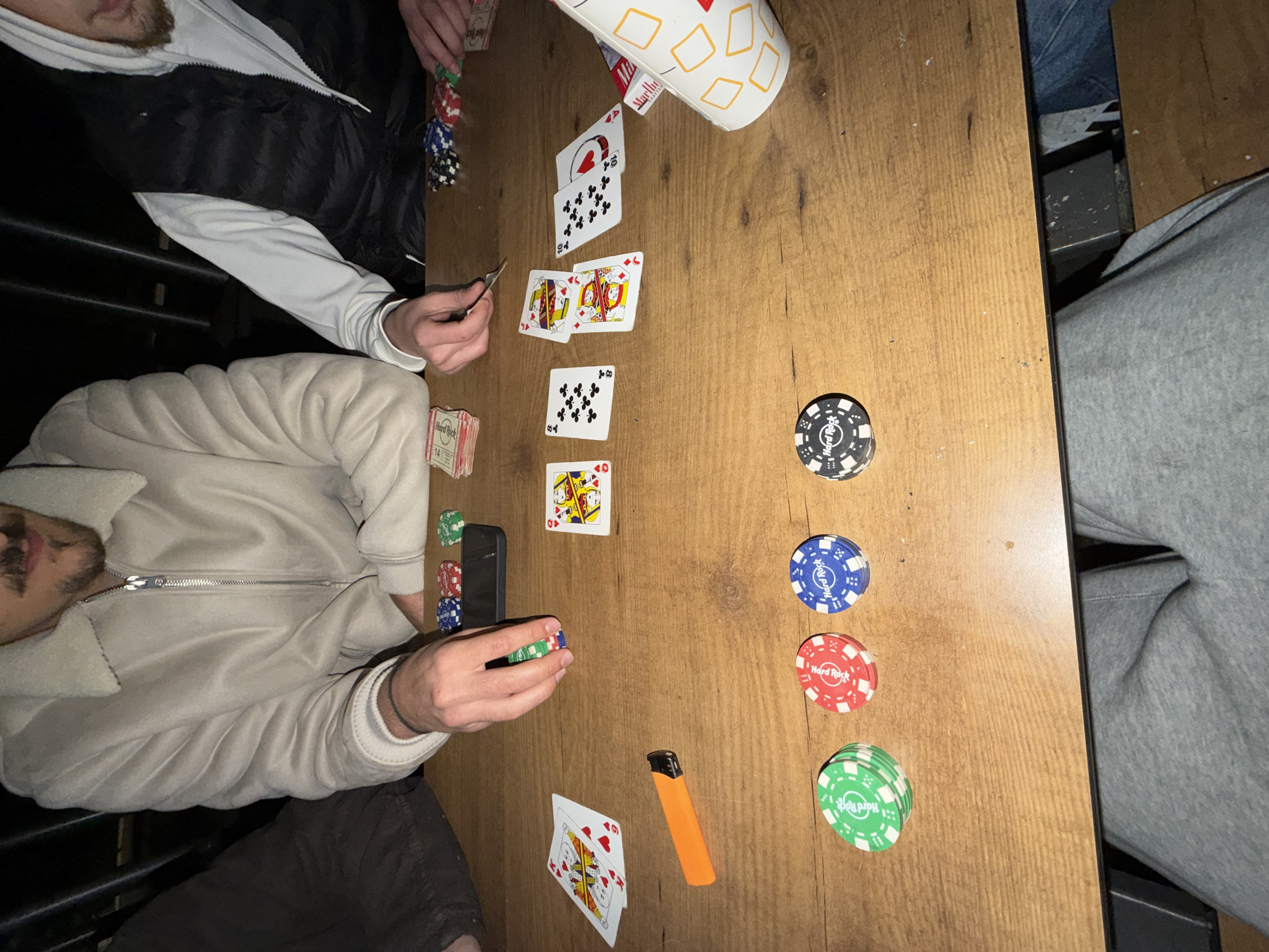 The image depicts a poker game in progress. Two individuals are visible, one wearing a white jacket and another wearing a black and white jacket. They are sitting at a wooden table with playing cards, poker chips, and cash visible. The table also contains a cell phone, a lighter, and a cup. The poker chips are branded "Hard Rock".
The location is not identifiable from the image. Therefore, identifying that the game is in Vila Real, Portugal is not possible.