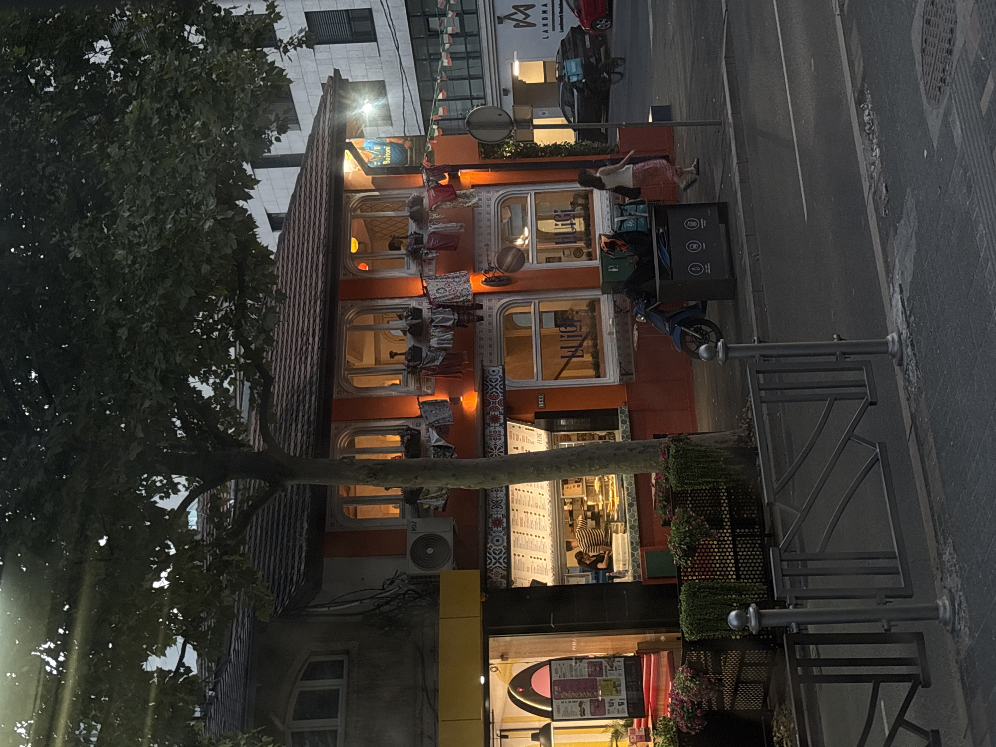 The image depicts a street scene in Belgrade, Serbia. The foreground features a dark grey metal fence and a paved sidewalk. Beyond the fence, a street is visible with a white line marking a lane.
The main focus is a two-story building with an orange facade and a brown tiled roof. The building houses a business, possibly a restaurant or cafe, with large windows displaying menus and food items. Two individuals are visible behind a counter inside, preparing food. There are several windows on the upper floor, some of which are illuminated from within. Laundry is hanging on a line outside the upper windows. A large tree with dark green foliage partially obscures the building.
To the right of the orange building, a modern white building rises. A sign with the letters "LM" and the word "LANDMARK" is visible on the white building. There are parked cars on the street, including a black car with a delivery box on the back and a red car. A blue scooter is parked near a green trash bin with logos on it. A woman in a light-colored top and a pink skirt is walking past the trash bin.
The lighting suggests it is either late afternoon or early evening, with some artificial lights illuminating the building's interior and exterior. The overall atmosphere is urban, with elements of commerce and residential buildings.