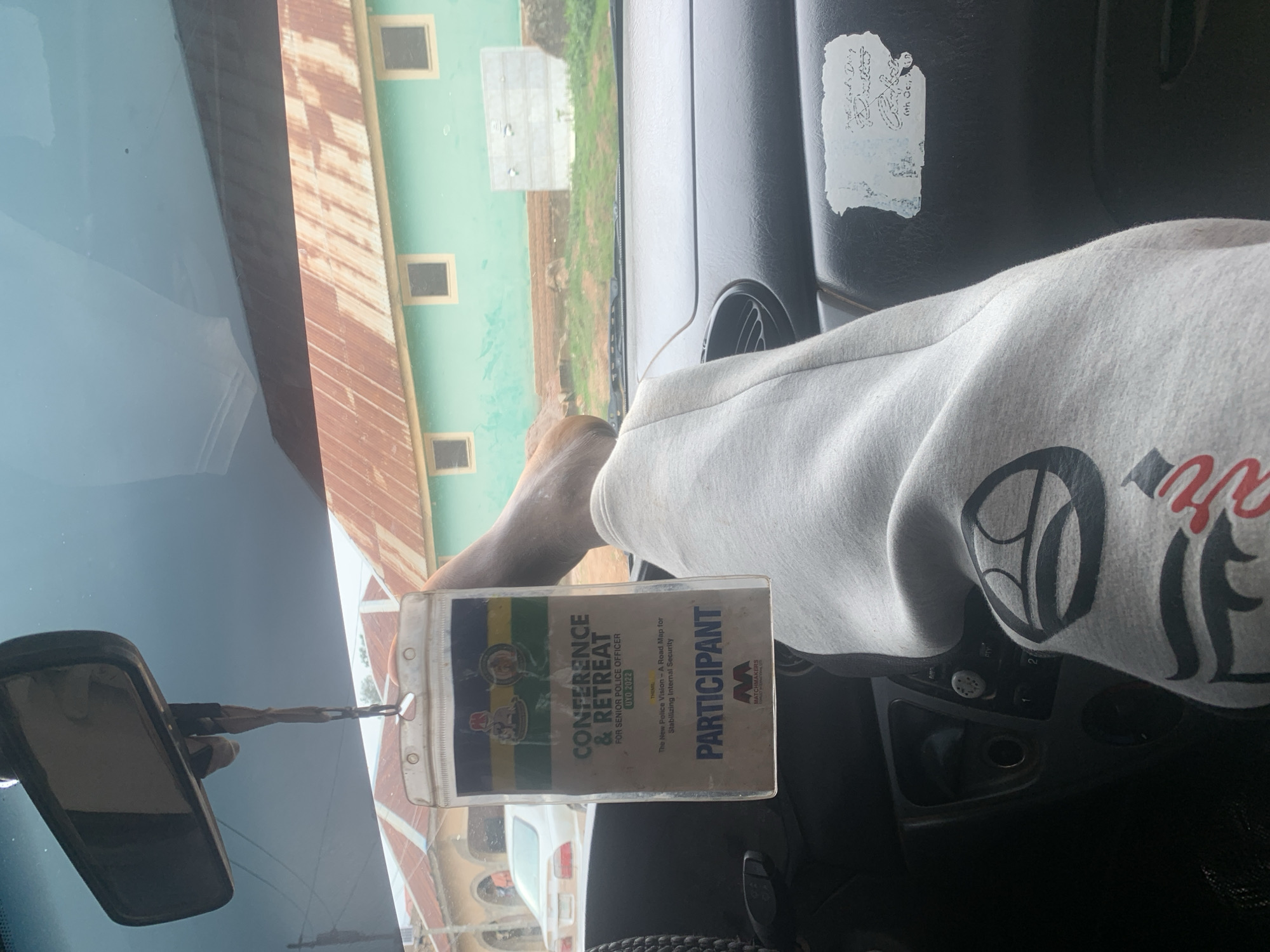 This image provides an interior view of a vehicle in Jos, Nigeria, with a focus on a participant's badge hanging from the rearview mirror. The badge identifies the holder as a participant in a "Conference & Retreat for Senior Police Officer UYO 2022," with the theme "The New Police Vision - A Road Map for Stabilizing Internal Security." A portion of a person's leg, clad in grey sweatpants with a dark graphic design, is visible in the foreground, resting against the dashboard. Through the windshield, a light green building with a corrugated iron roof is visible, suggesting an outdoor environment typical of Jos. The scene appears to be a still moment during a journey or waiting period, with the badge prominently displayed as evidence of the individual's engagement in the police conference.